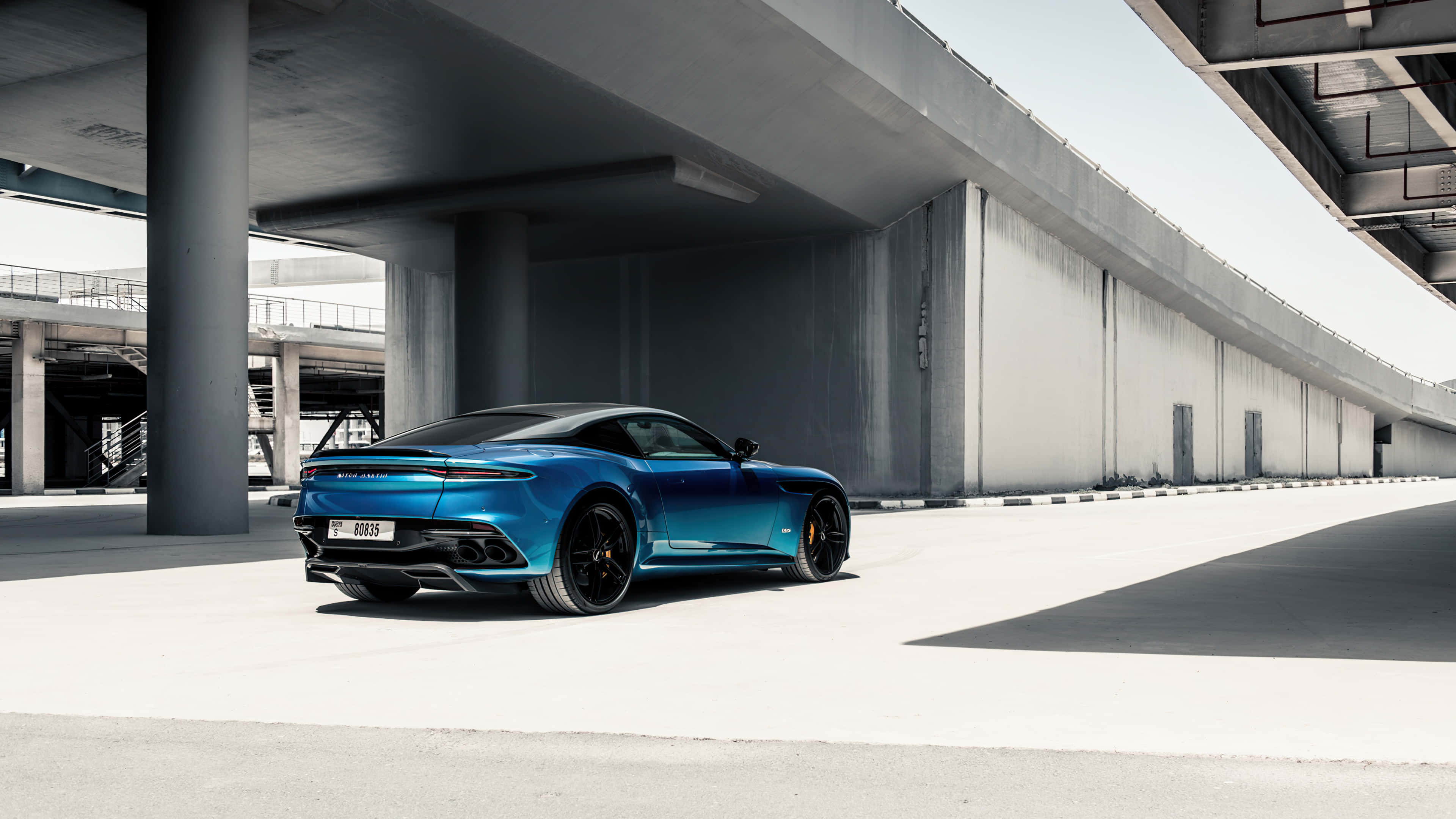 Aston Martin Dbs Superleggera 桌布 壁紙 — 一架時尚的藍色跑車停泊在現代混凝土天橋下，帶有銳利陰影和極簡主義建築風格。