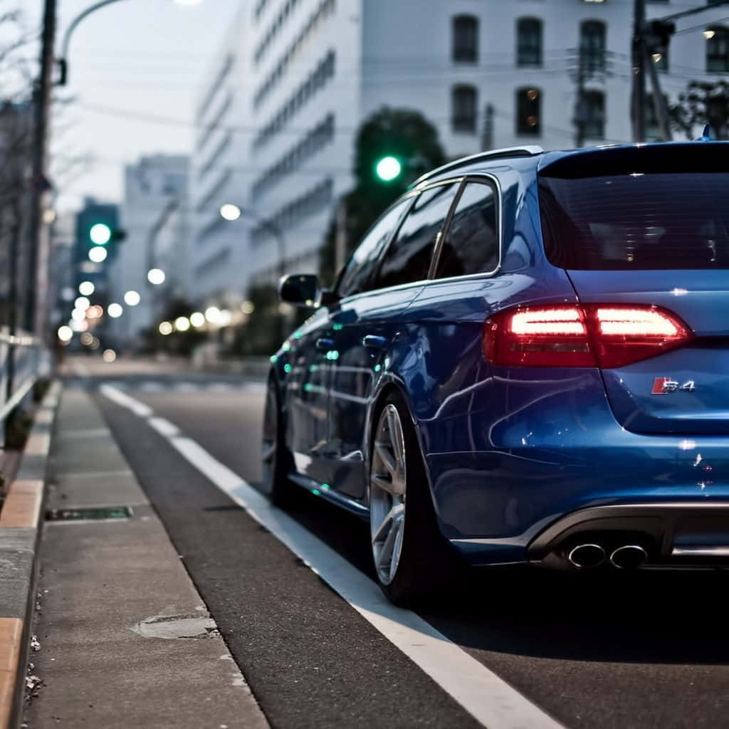 Audi S4 桌布 壁紙 — 一輛時尚的藍色旅行車在黃昏時停泊在城市的街道上，尾燈發光，背景模糊的城市景觀。