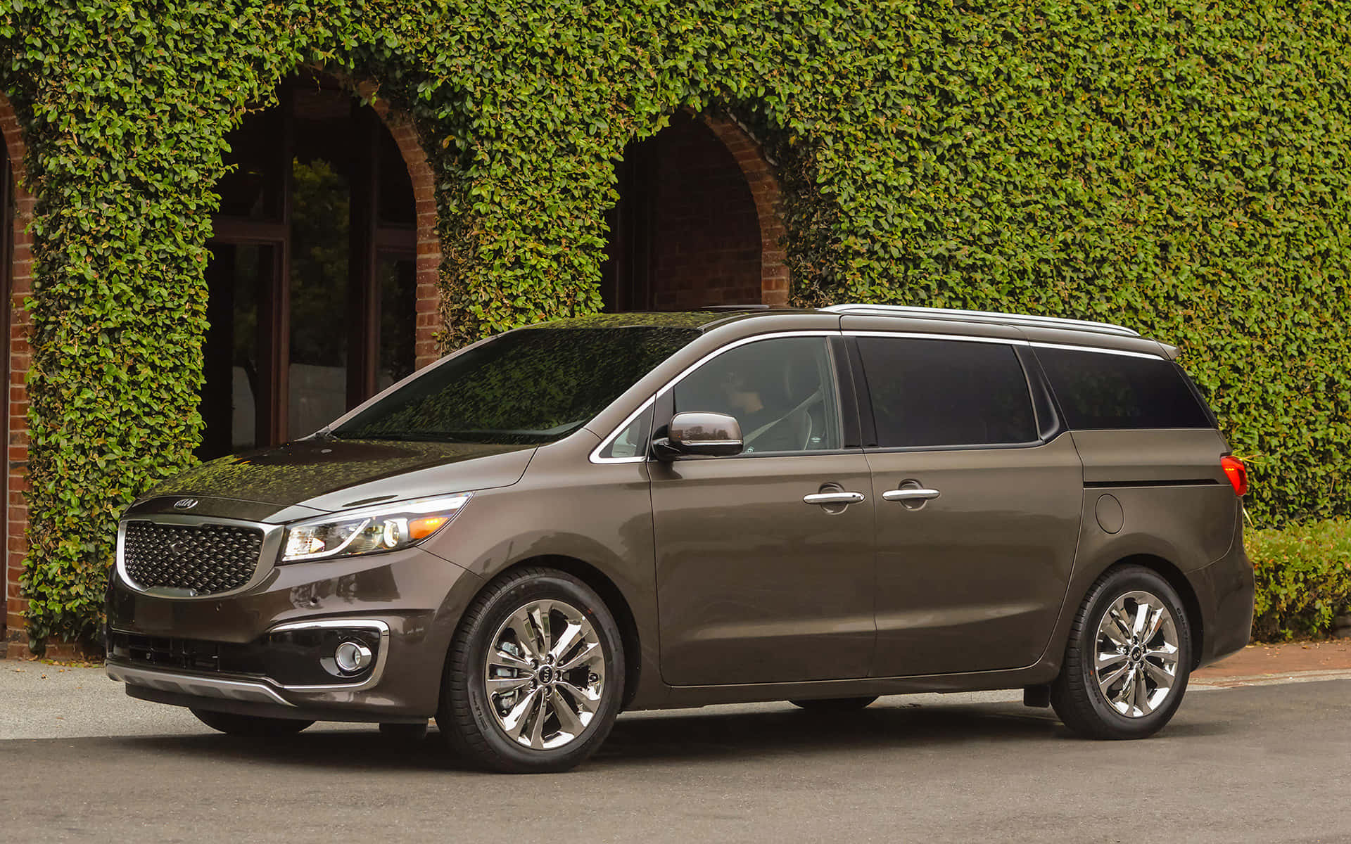 Kia Sedona 桌布 壁紙 — 一輛時尚的棕色Kia小型貨車停在佈滿茂密綠色常春藤的磚砌建築前。