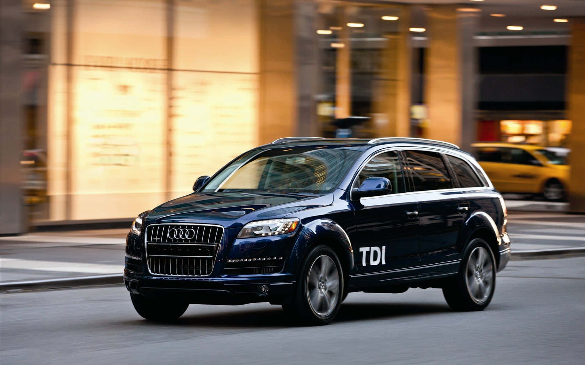 Audi Q7 桌布 壁紙 — 一輛時尚的深藍色Audi Q7 TDI SUV在城市街道上行駛，背景是模糊的商店櫥窗和一輛黃色的士。
