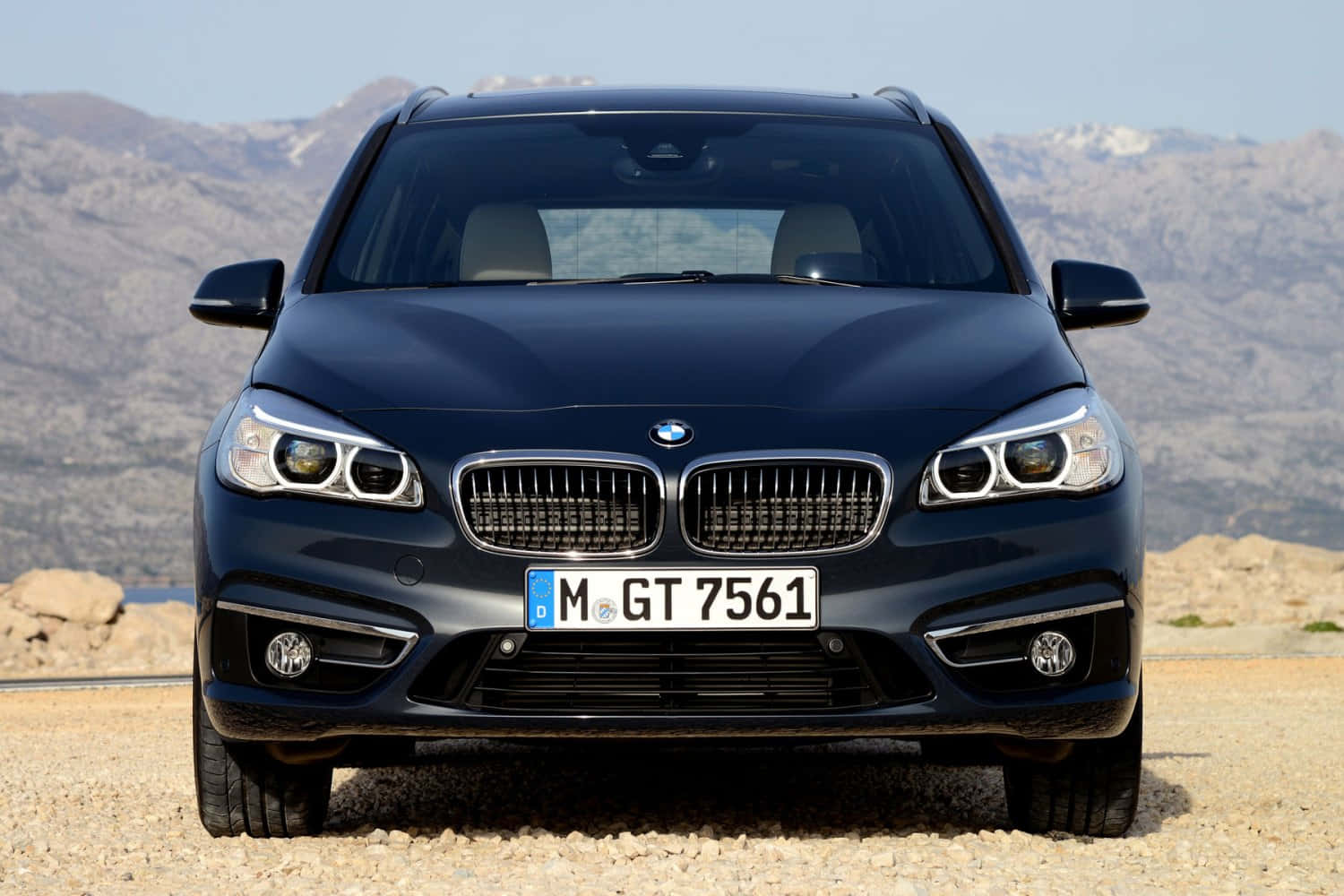 Bmw 2 Series 桌布 壁紙 — 一輛時尚深藍色寶馬SUV在碎石路上向前行駛，背景是晴朗天空下的崎嶇山脈。