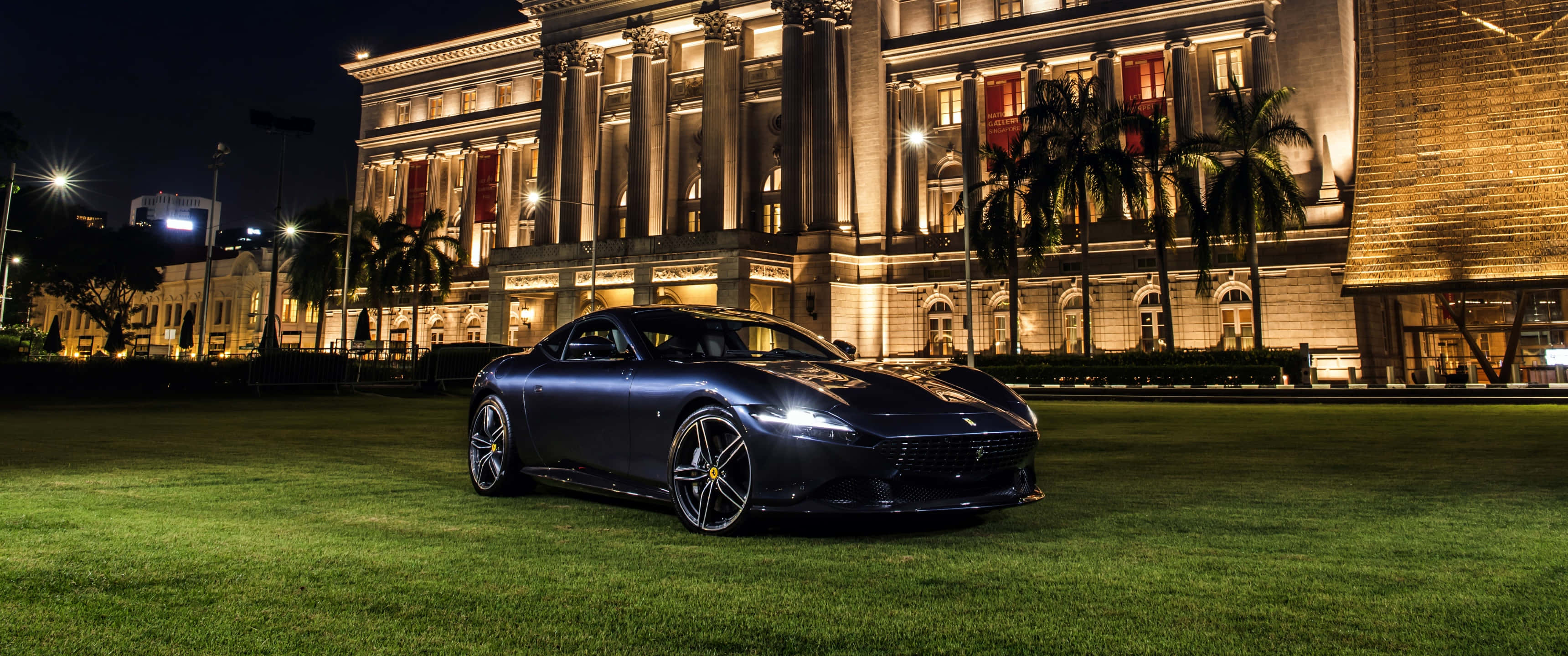 Ferrari Roma 桌布 壁紙 — 一輛時尚的深藍色法拉利跑車在夜燈下閃閃發光，停在綠色草地上，前面是一座宏偉的照明新古典主義建築。
