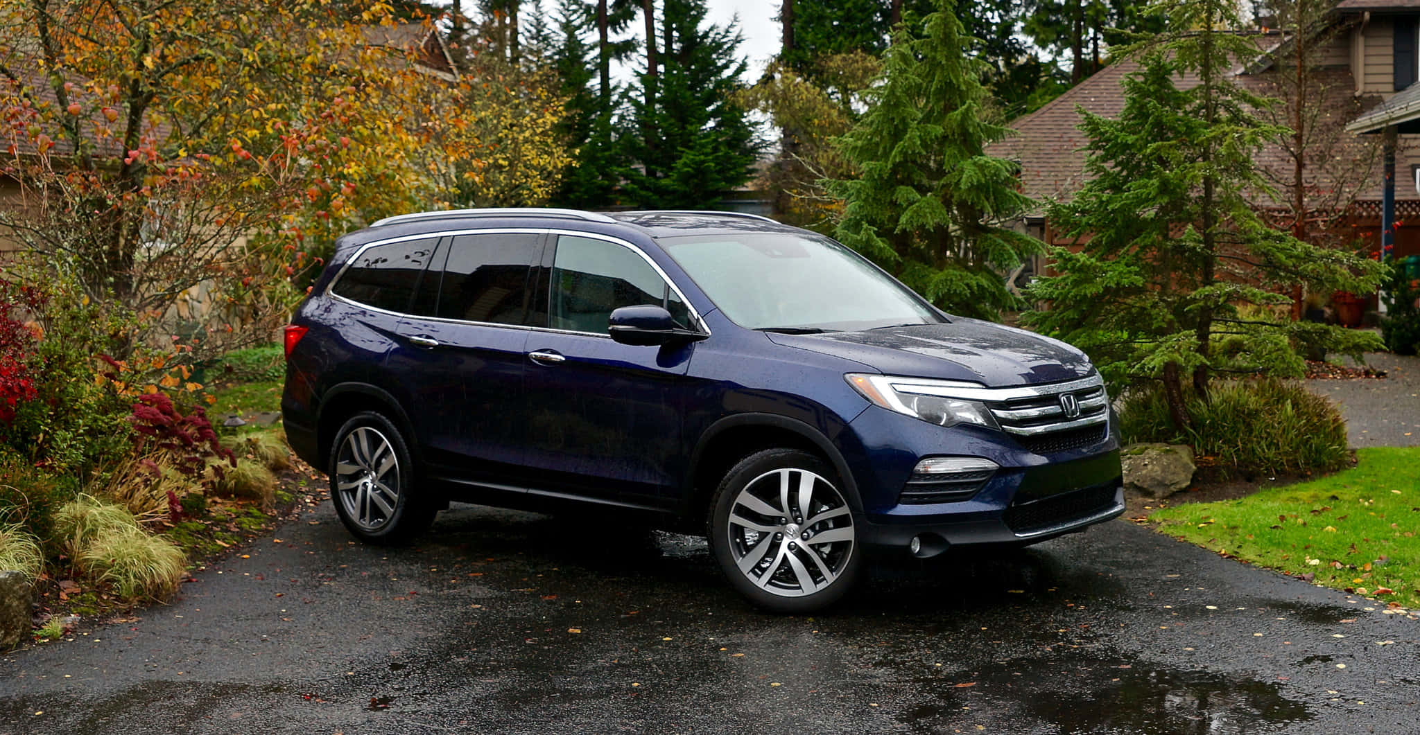 Honda Pilot 桌布 壁紙 — 一輛時尚的深藍色本田Pilot SUV停泊在濕漉漉的車道上，周圍環繞著秋葉和常綠樹木。