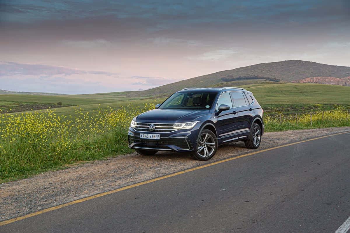 Volkswagen Tiguan 桌布 壁紙 — 一架時尚的深藍色SUV停泊在鄉村道路上，旁邊是鮮豔的黃色野花，背景是陰鬱的黃昏天空。