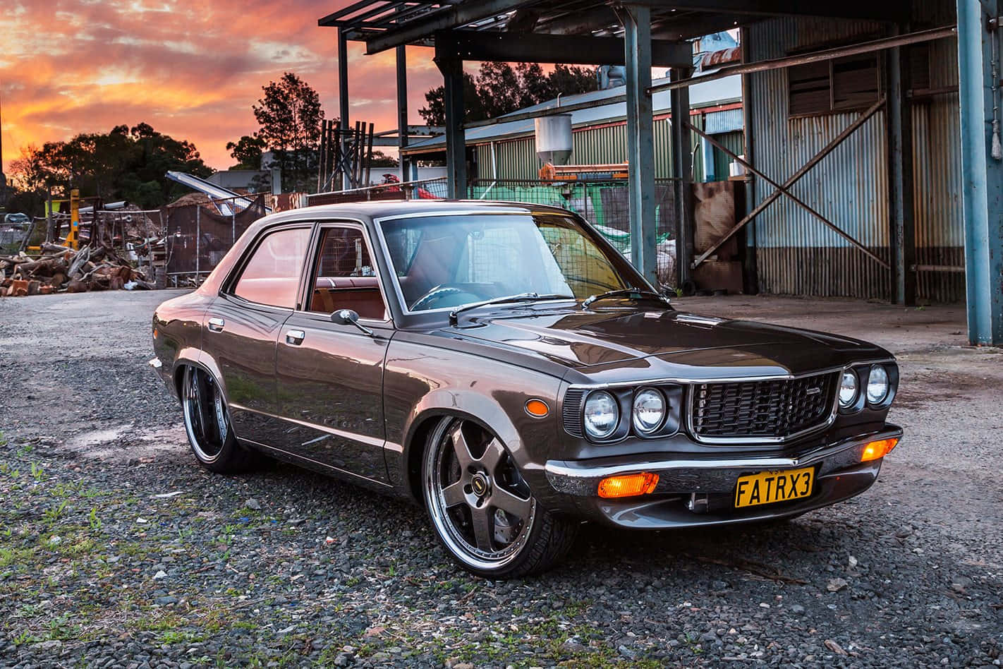 Mazda Rx-3 桌布 壁紙 — 一架時尚深棕色經典汽車配備定制輪圈，停在夕陽下生鏽工業建築前的碎石地上。
