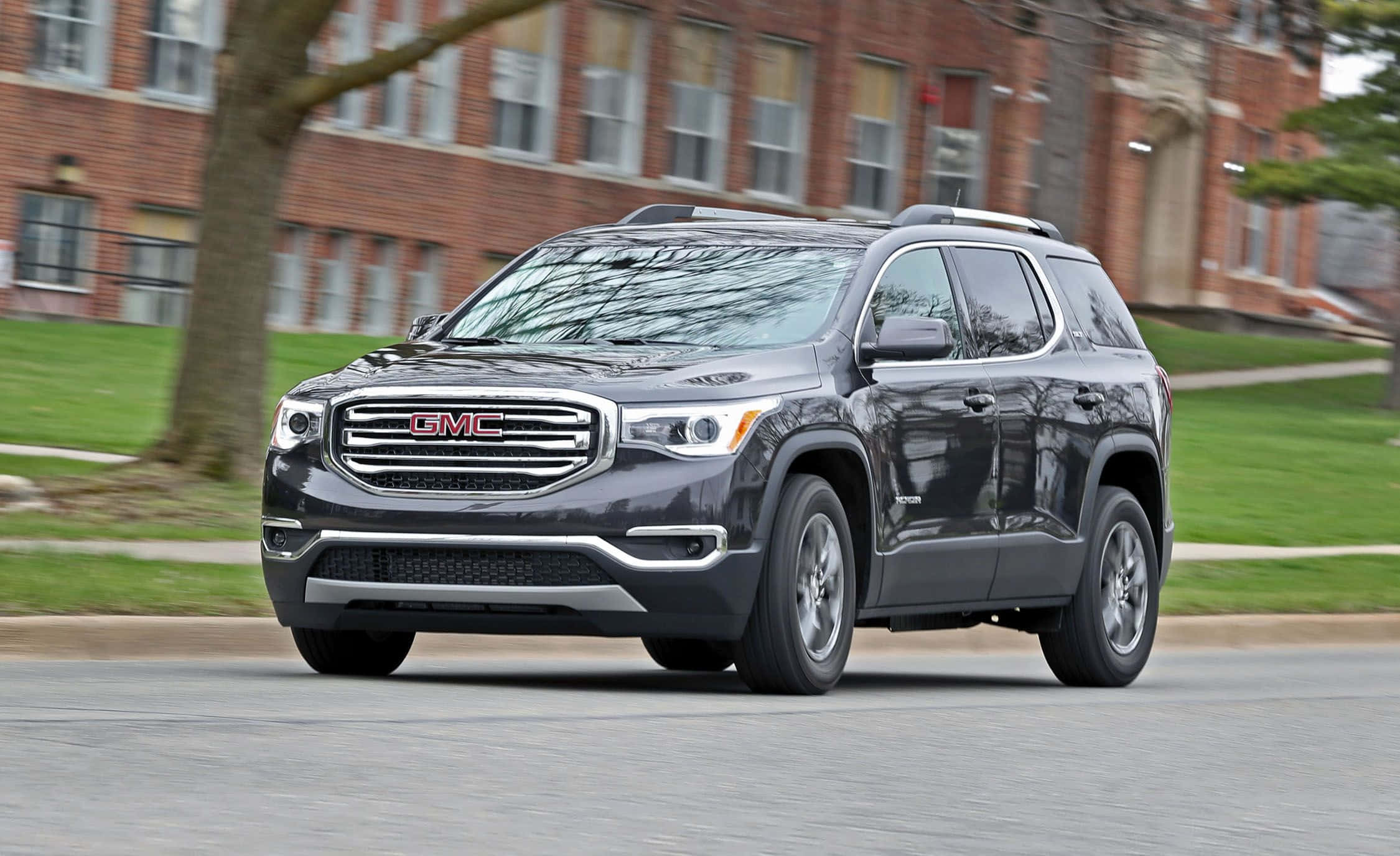 Gmc Acadia 桌布 壁紙 — 一輛時尚的深灰色 GMC Acadia SUV 在綠樹成蔭的城市街道上平穩行駛，背景是磚砌建築。