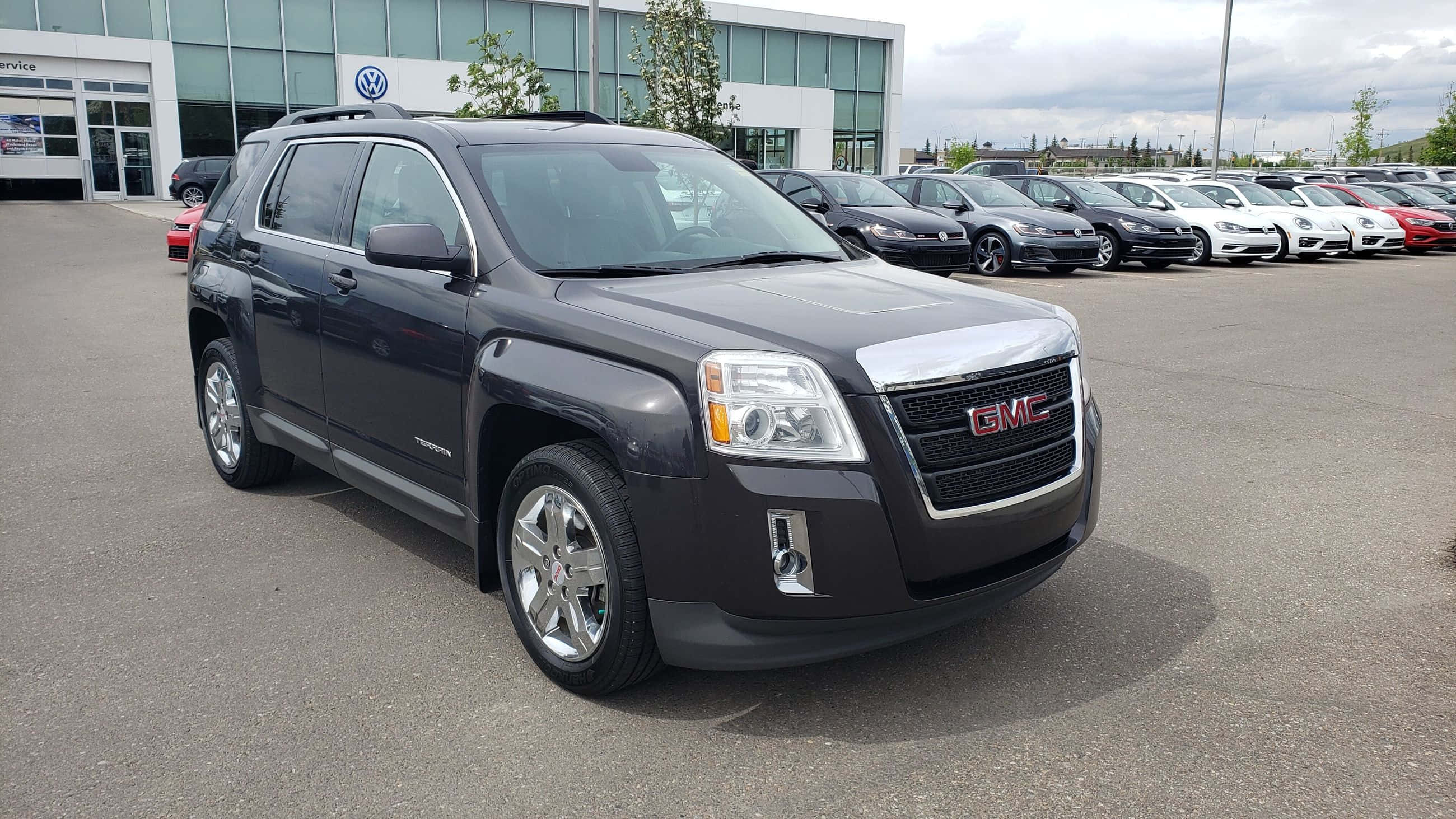 Gmc Terrain 桌布 壁紙 — 一輛時尚的深灰色 GMC Terrain SUV 停泊在經銷商場地，與其他車輛一起，天空多雲。