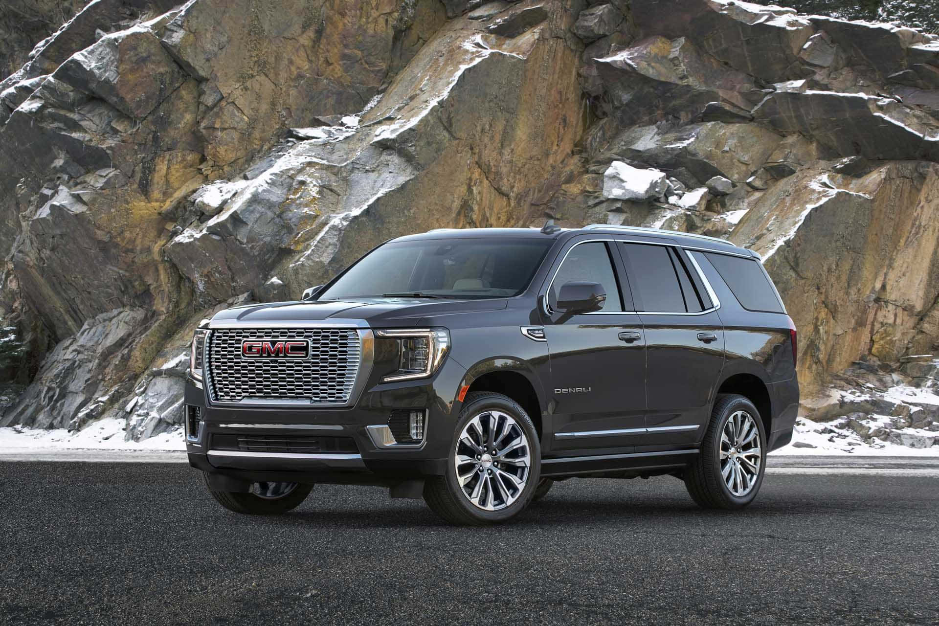 Gmc Yukon 桌布 壁紙 — 一架時尚深灰色GMC Yukon Denali SUV停泊在瀝青路上，前方是崎嶇積雪的岩石懸崖面。