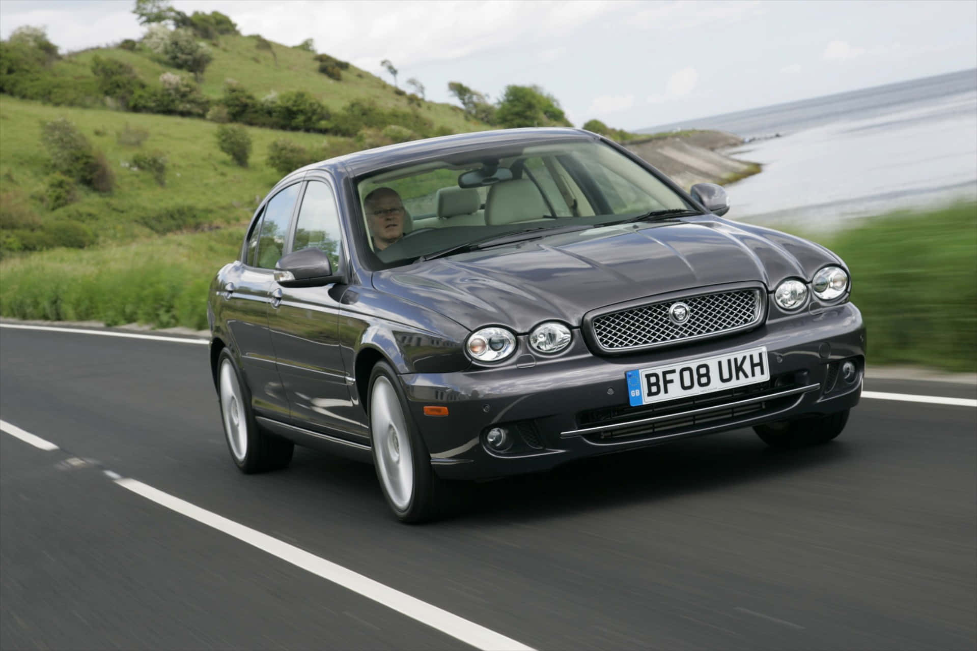 Jaguar X-type 桌布 壁紙 — 一輛時尚的深灰色積架房車沿著海岸公路疾馳，背景是綠色山丘和海洋。