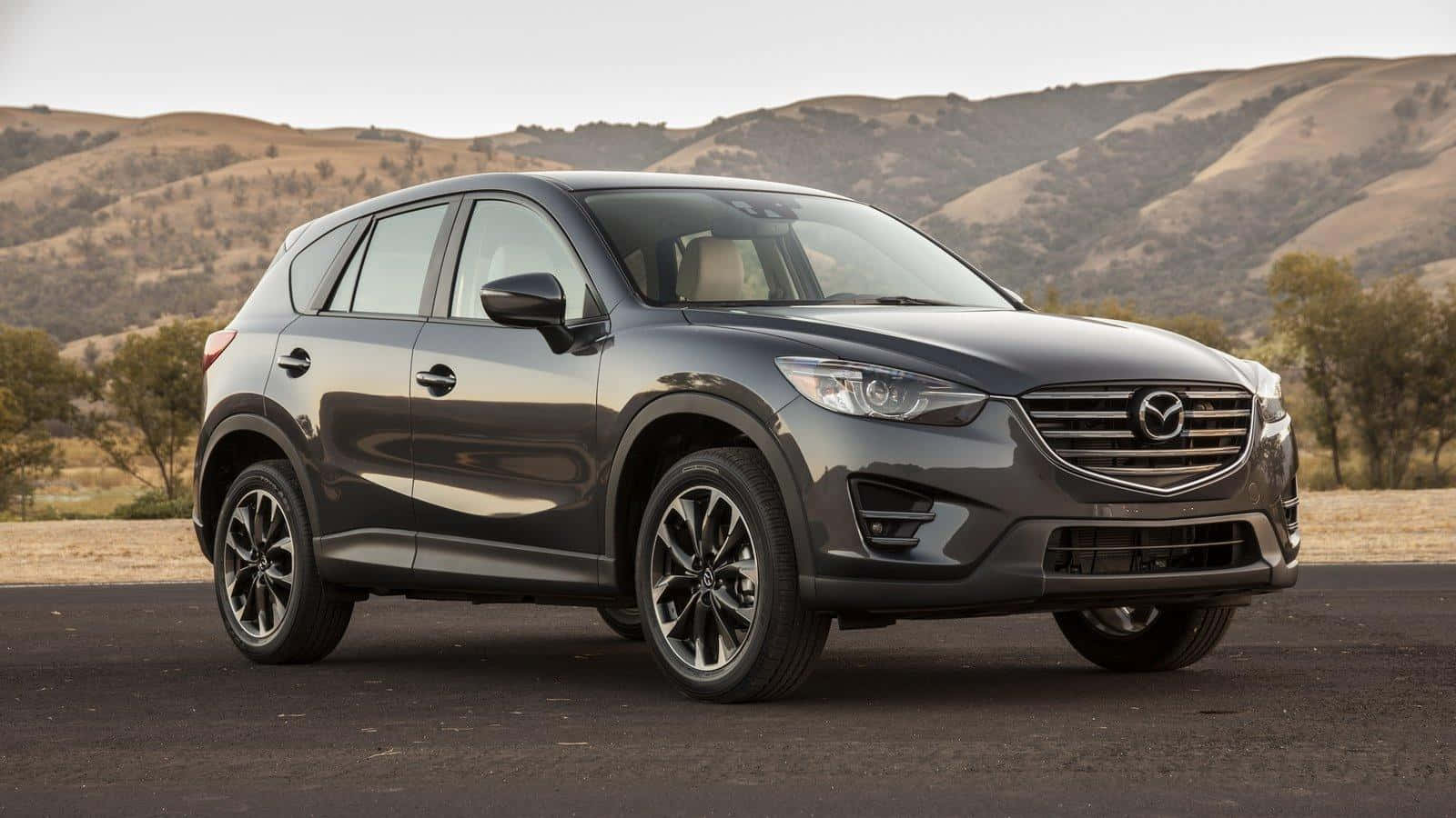 Mazda Cx-5 桌布 壁紙 — 一輛時尚的深灰色Mazda CX-5 SUV停在瀝青路上，背景是起伏的乾燥山丘和稀疏樹木，在柔和日光下。