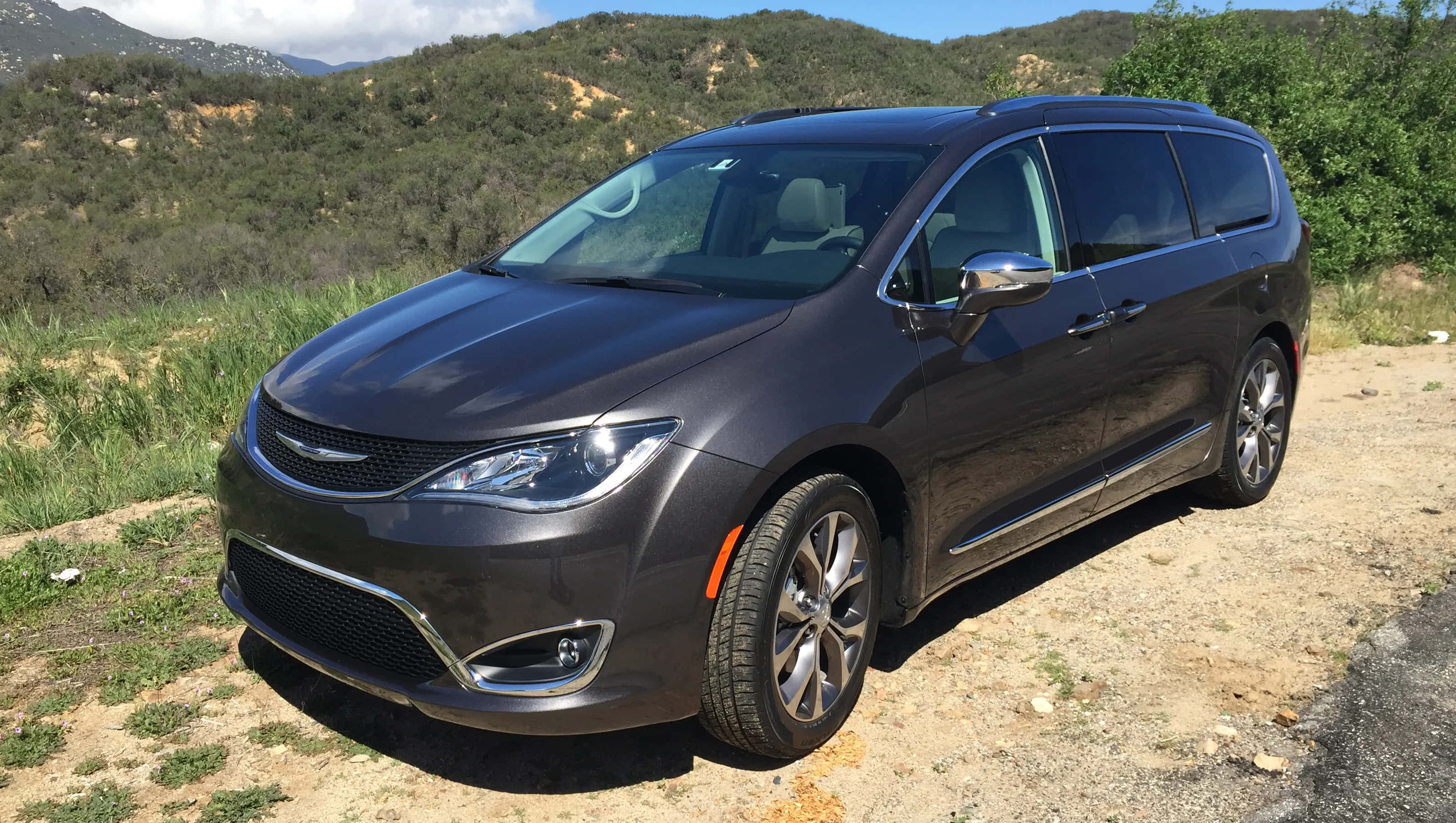 Chrysler Pacifica 桌布 壁紙 — 一架時尚深灰色小型客車停泊在泥路上，周圍是起伏的綠色山丘和散落的樹木，頭頂是明亮的藍天。