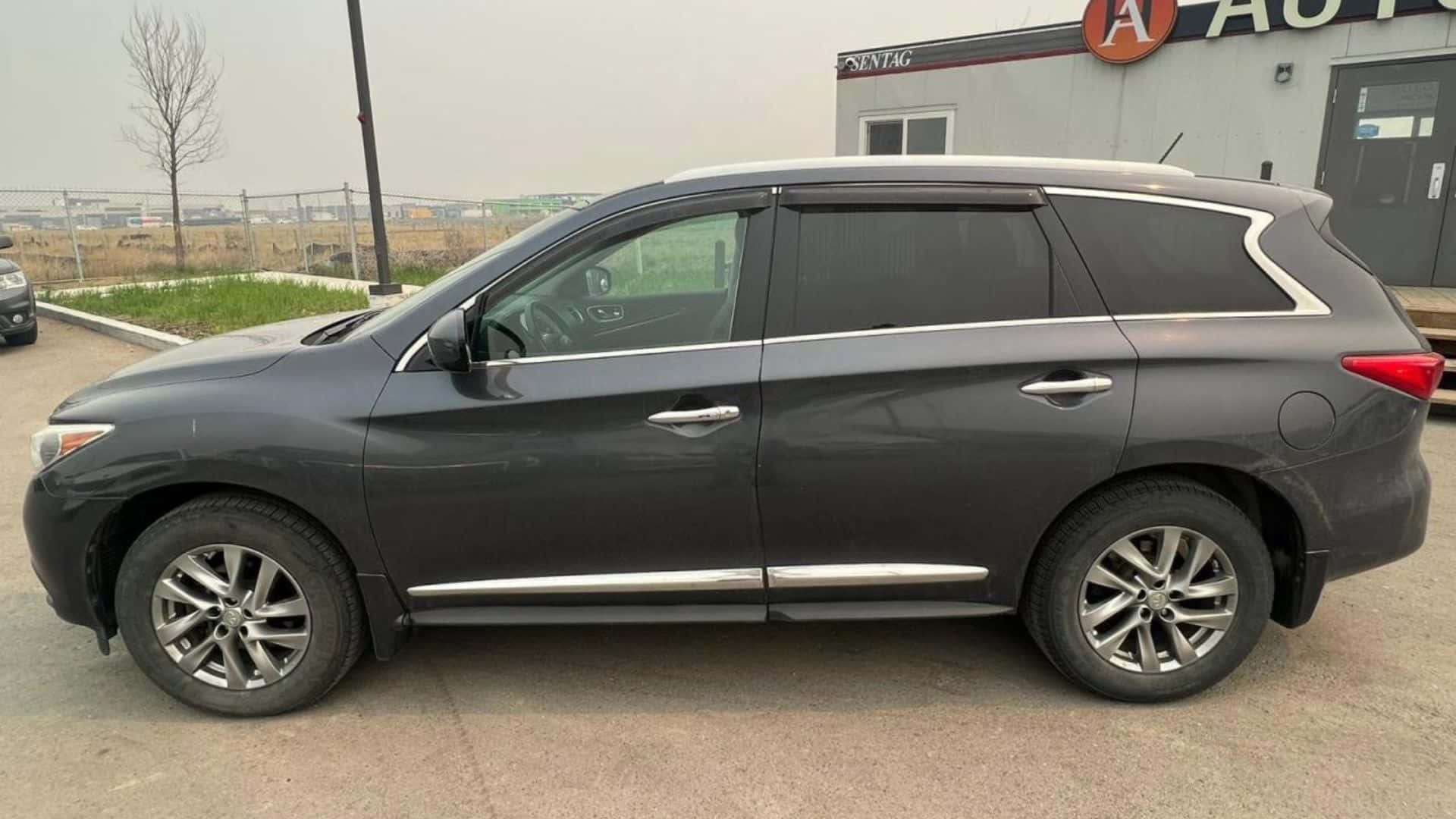 Infiniti Jx35 桌布 壁紙 — 一輛時尚的深灰色SUV停泊在經銷商外，背景有光禿的樹木和陰天。