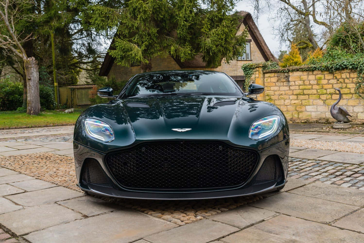 Aston Martin Dbs Superleggera 桌布 壁紙 — 一輛時尚的深綠色 Aston Martin 跑車在陰天天空下閃閃發光，停在石頭車道上，旁邊是質樸的石牆和樹木。