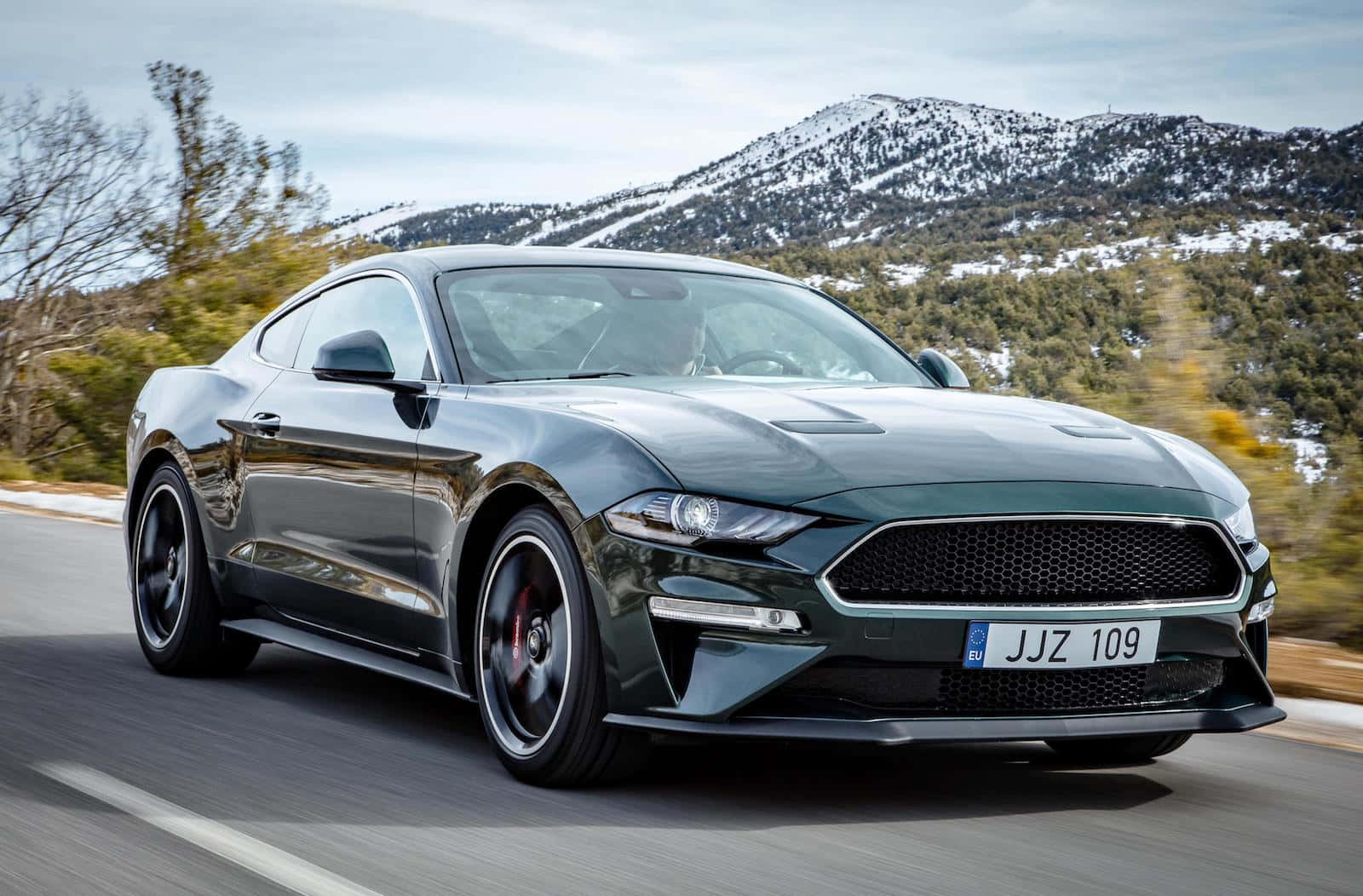 Ford Mustang Bullitt 桌布 壁紙 — 一架流線型深綠色福特野馬在雪山頂和秋樹背景下沿山路飛馳。