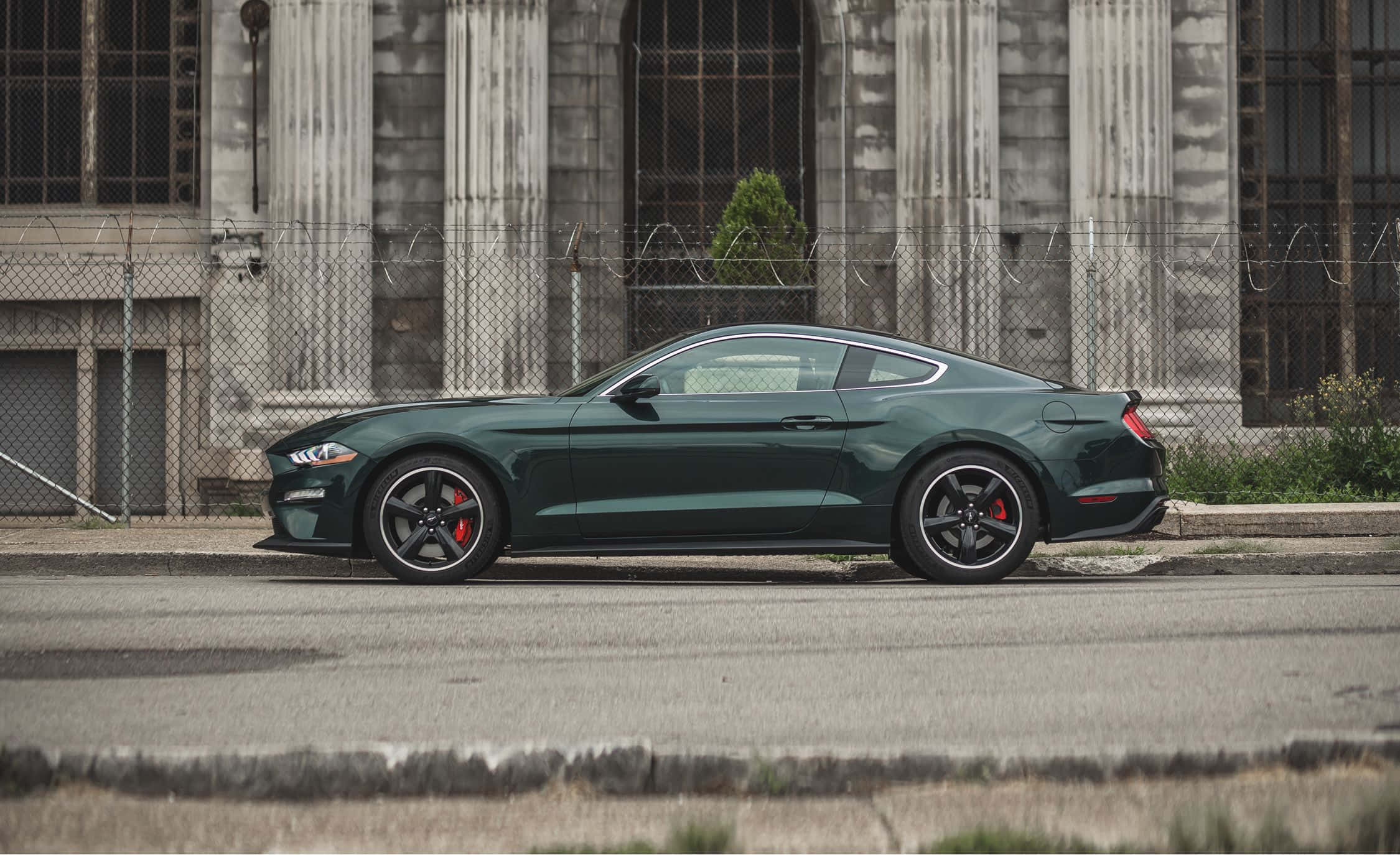 Ford Mustang Bullitt 桌布 壁紙 — 一架時尚的深綠色福特野馬跑車停泊在佈滿灰塵的城市街道上，旁邊是鐵絲網圍欄和風化的石建築。