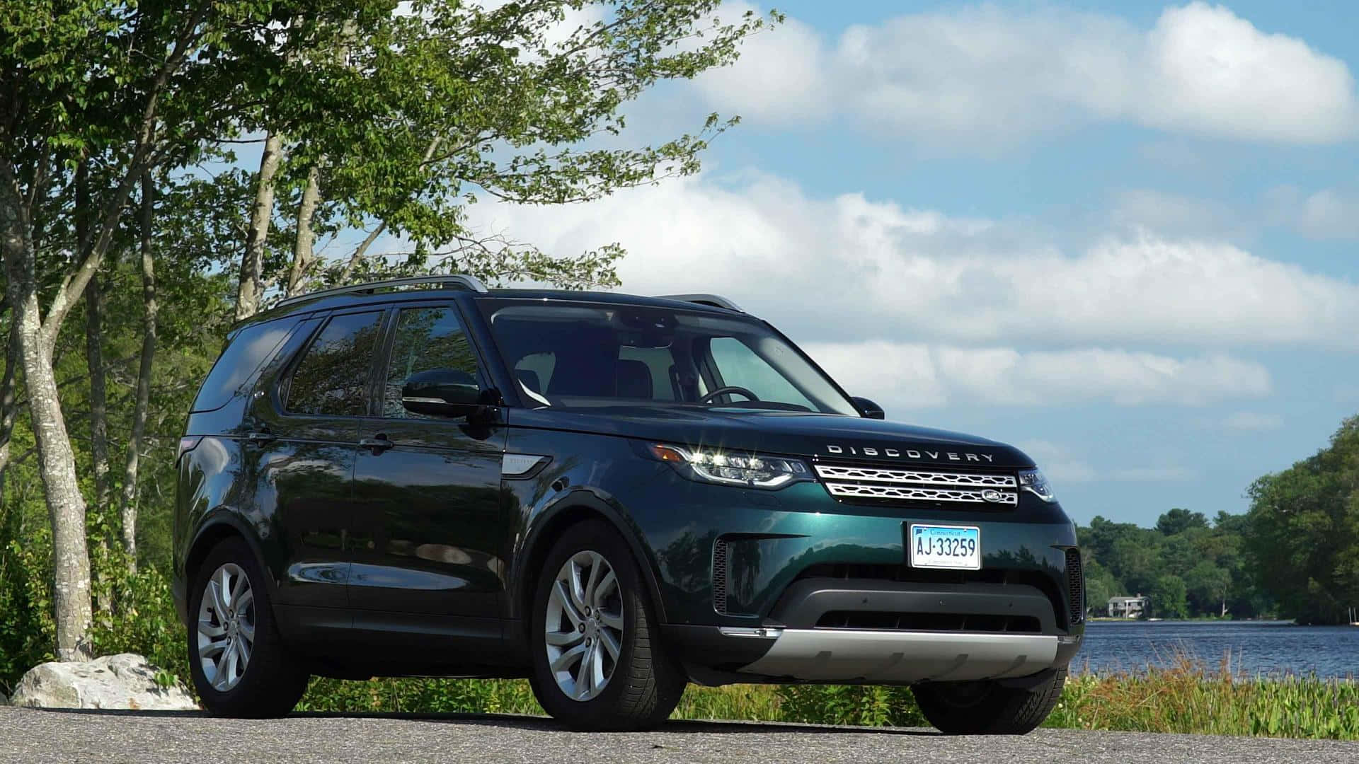Land Rover Discovery 桌布 壁紙 — 一輛時尚的深綠色 Land Rover Discovery 停泊在碎石上，旁邊是一個寧靜的湖泊，天空部分多雲。