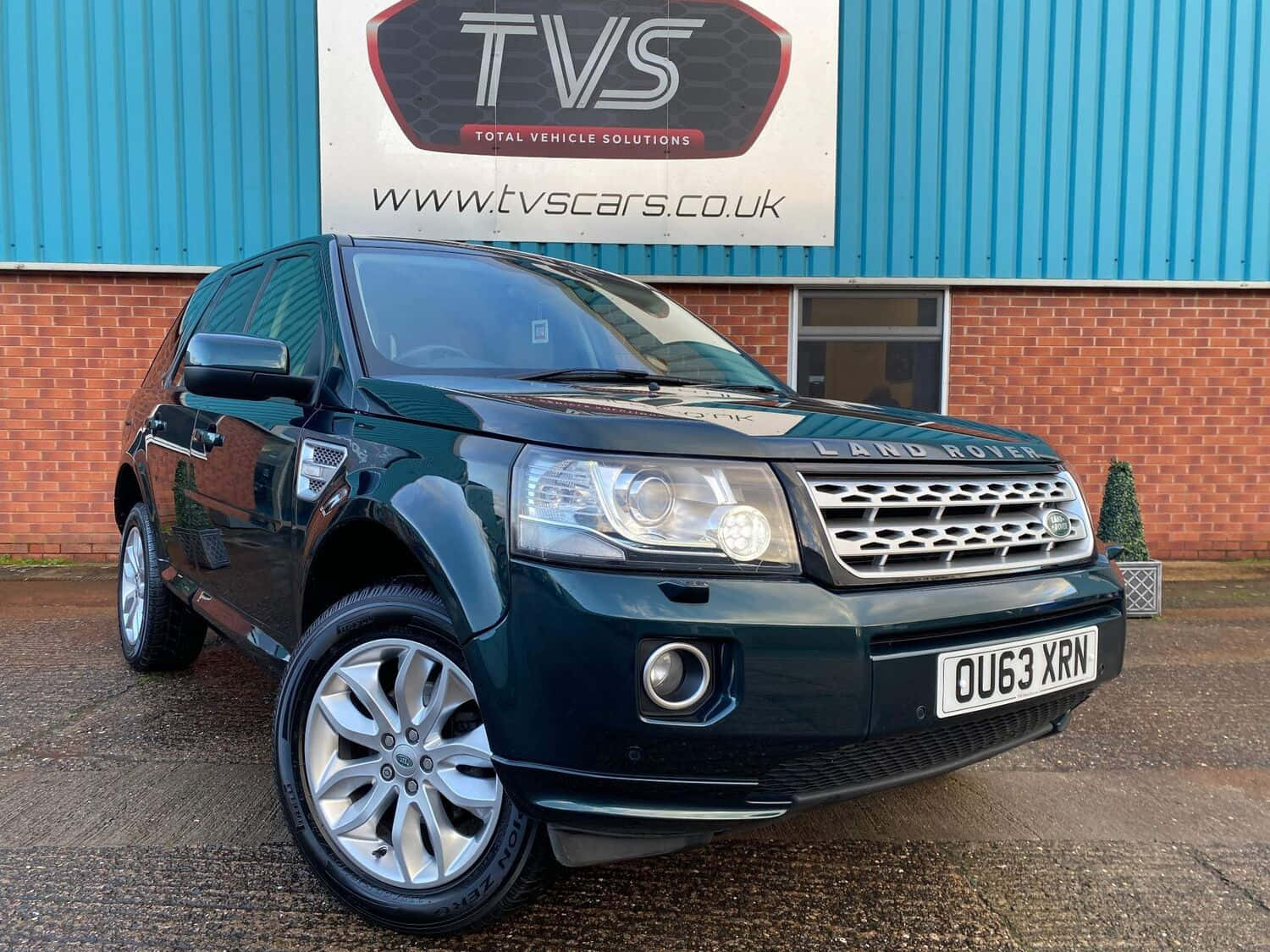 Land Rover Freelander 桌布 壁紙 — 一輛時尚的深綠色 Land Rover Freelander 停在碎石路上，前面是藍色波紋金屬和磚砌建築，帶有 TVS 標誌。