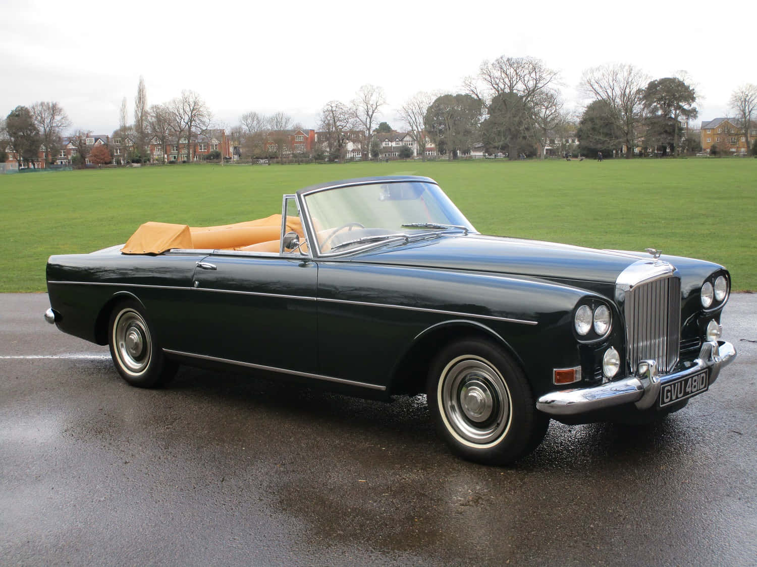 Bentley S3 桌布 壁紙 — 一架時尚深綠色復古開篷車，配米色內飾，停泊在潮濕路面上，旁邊是廣闊的綠色田野，天空多雲。