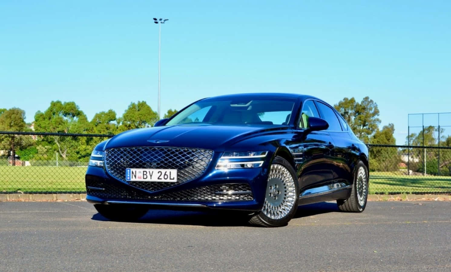 Genesis G70 桌布 壁紙 — 一輛時尚的深藍色豪華轎車在明亮的陽光下閃閃發光，停在瀝青路上，背景有綠樹和圍欄。