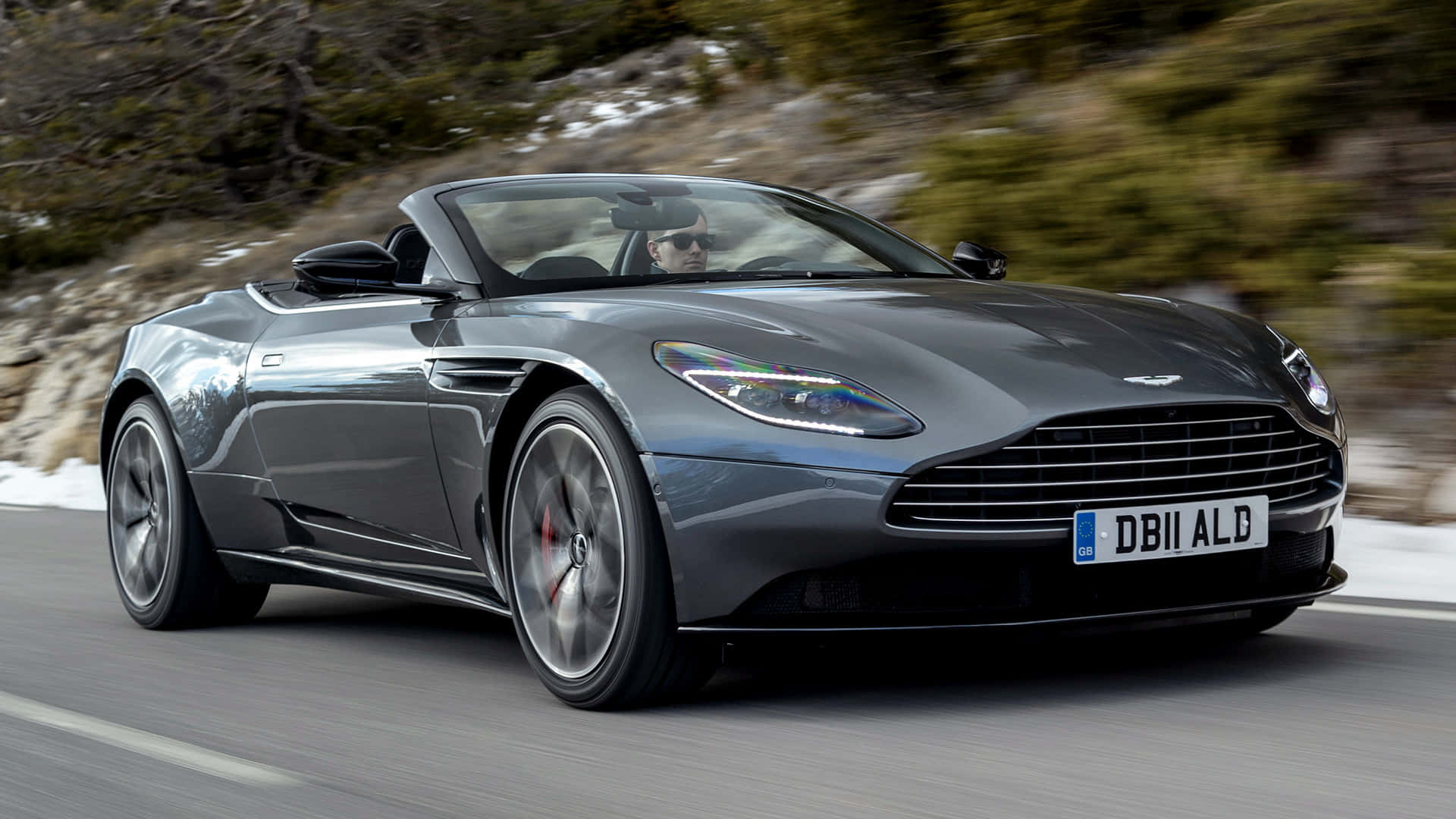 Aston Martin Db11 桌布 壁紙 — 一輛時尚的灰色 Aston Martin DB11 開篷跑車沿著蜿蜒的山路疾馳，兩旁是白雪覆蓋的懸崖和模糊的綠色植物。