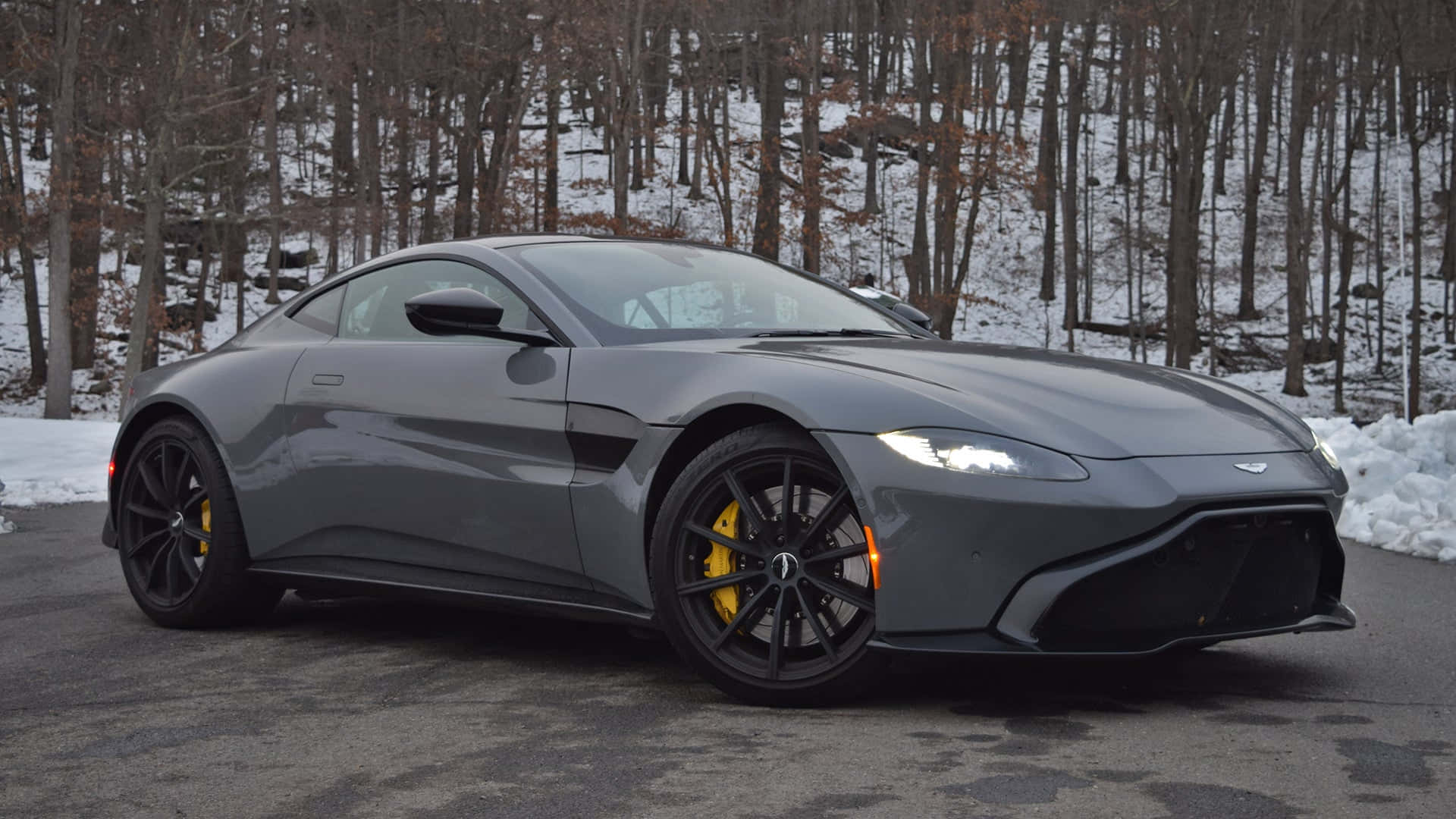Aston Martin Vantage 桌布 壁紙 — 一架時尚灰色 Aston Martin 跑車，配備黃色制動卡鉗，停泊在積雪的森林道路上。