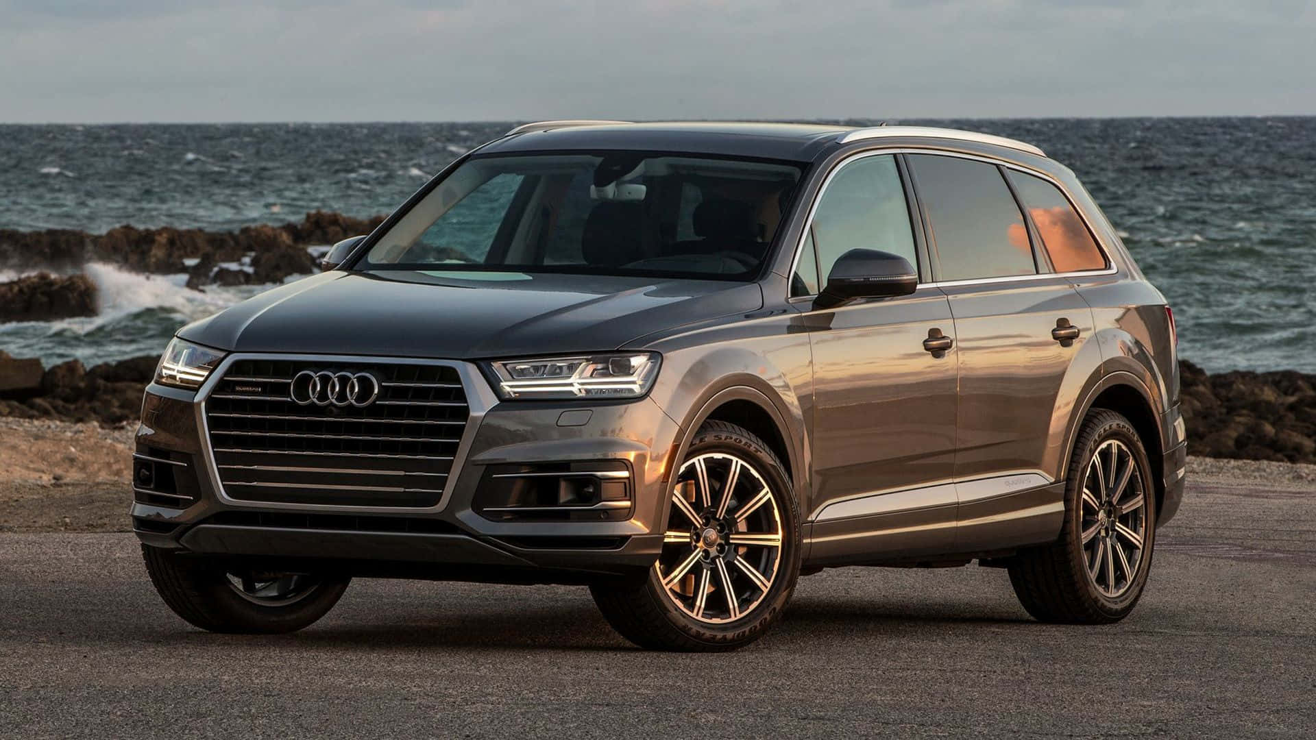 Audi Q7 桌布 壁紙 — 一架時尚灰色奧迪Q7 SUV停泊在海岸公路上，背後有海浪拍打岩石。