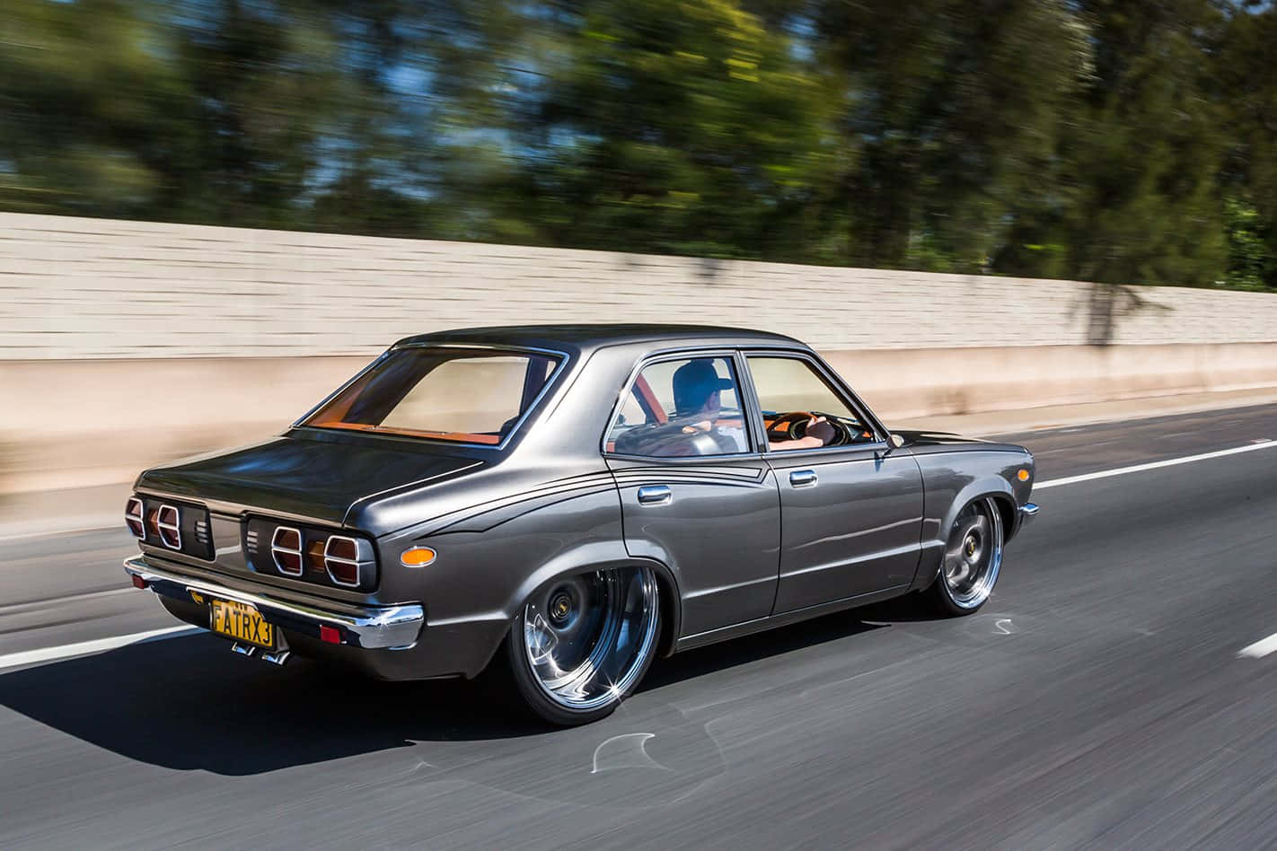 Mazda Rx-3 桌布 壁紙 — 一輛時尚的灰色經典汽車配備定制輪圈，在陽光明媚的高速公路上疾馳，背景是模糊的綠色樹木和混凝土屏障。