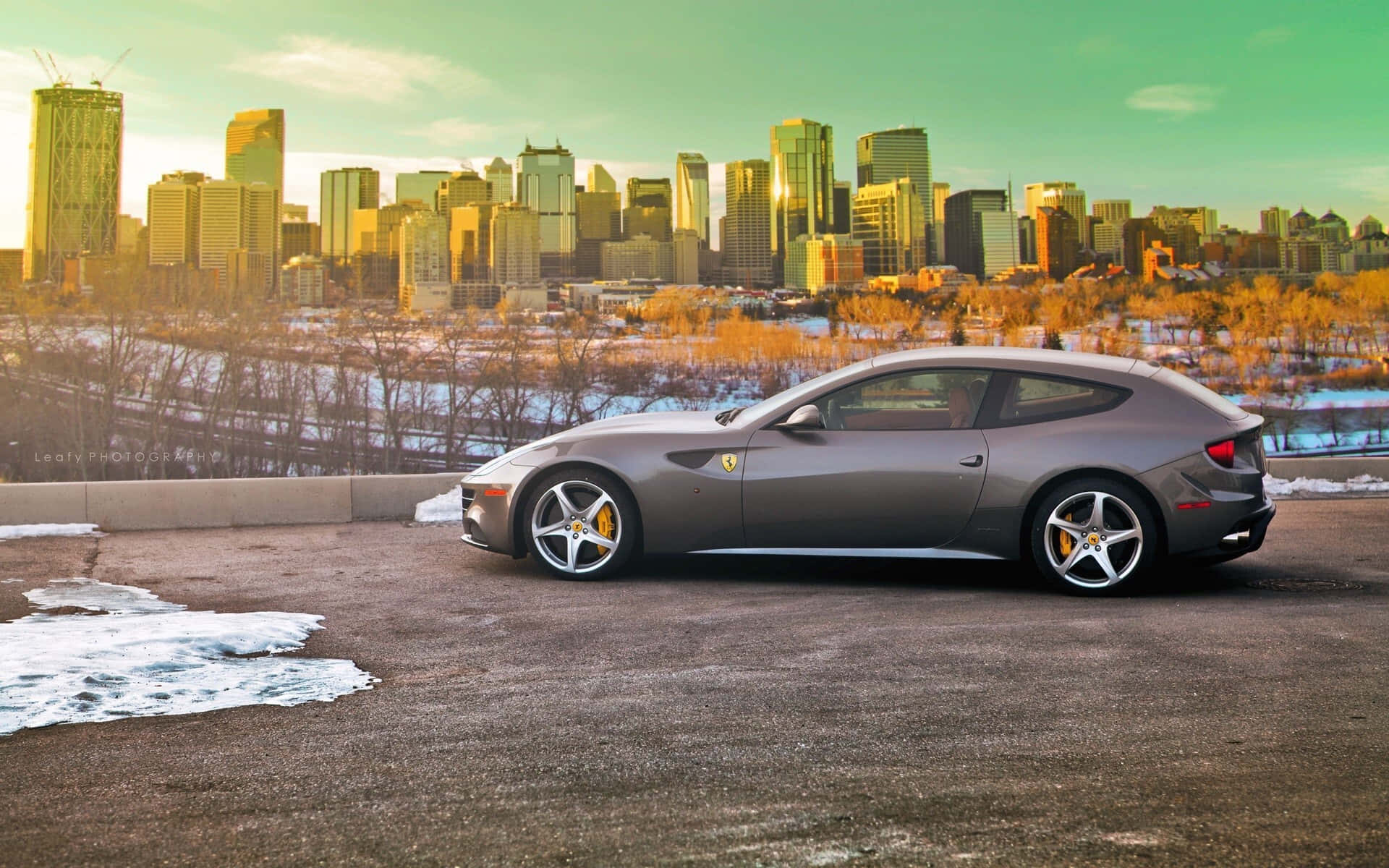 Ferrari Ff 桌布 壁紙 — 一架時尚的灰色法拉利停泊在雪景觀景點，背景是日落時金黃色的城市天際線。