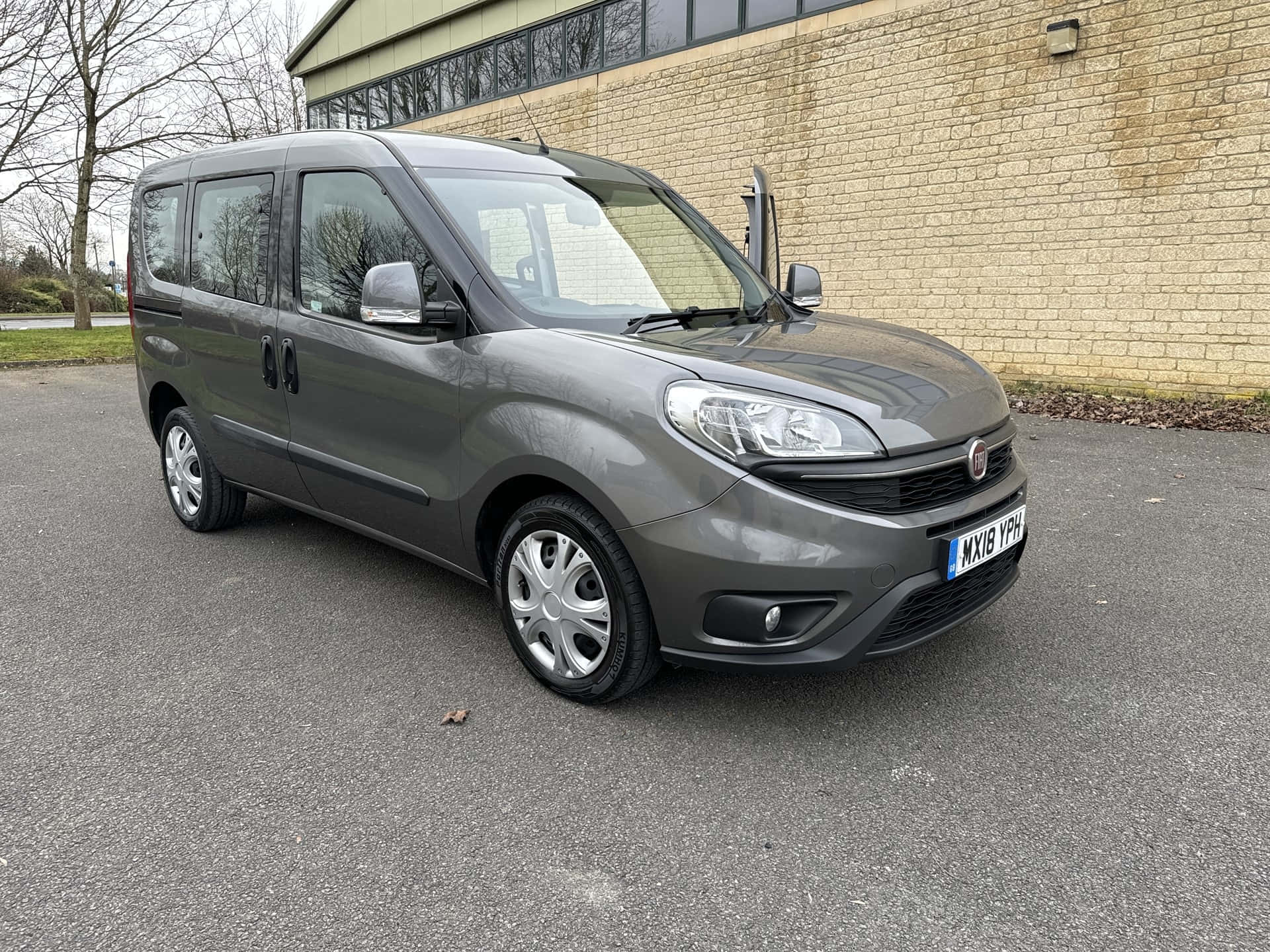  壁紙 — 一架時尚的灰色 Fiat Fiorino 客貨車停泊在瀝青路上，旁邊是米色磚砌建築，背景有光禿禿的樹木。