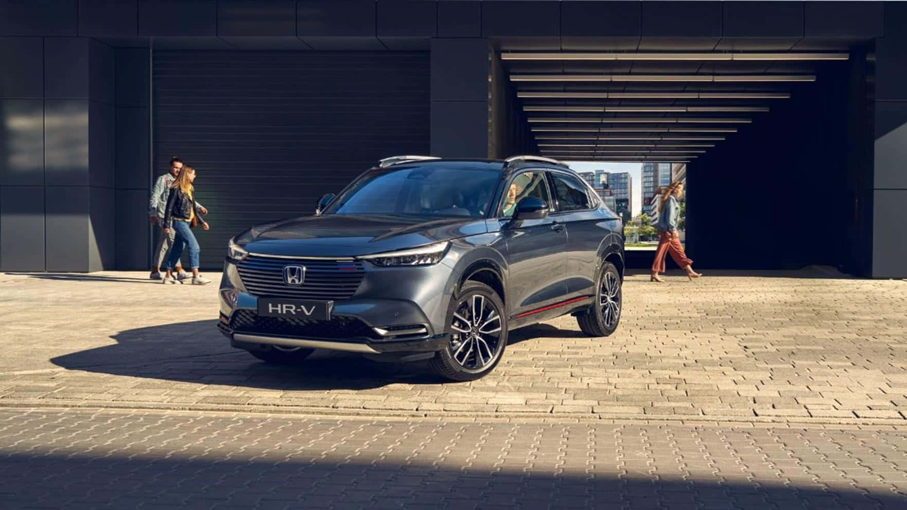 Honda Hr-v 桌布 壁紙 — 一架時尚的灰色本田HR-V SUV停泊在陽光普照的磚砌廣場上，人們在附近的現代建築結構下行走。