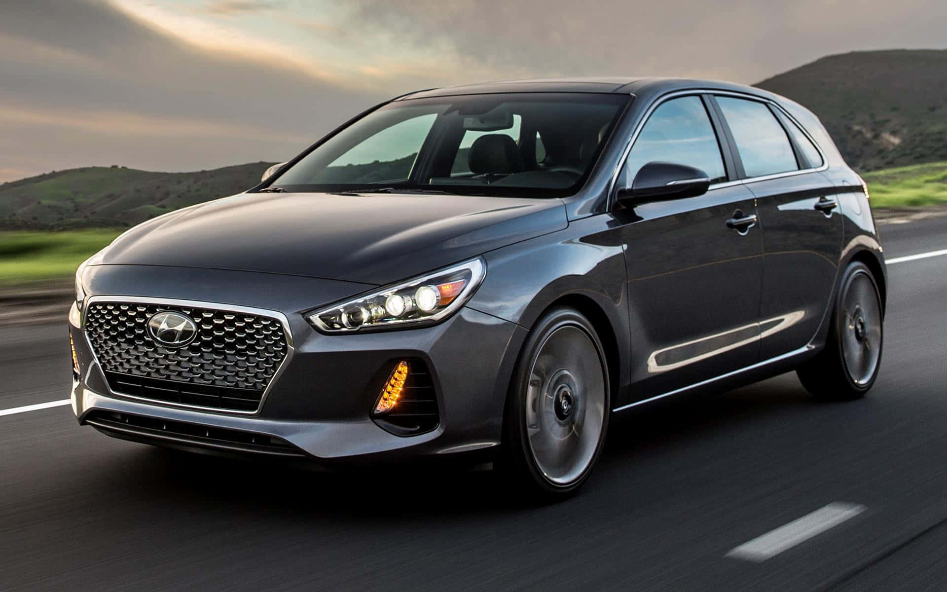 Hyundai Elantra 桌布 壁紙 — 一輛時尚的灰色現代掀背車在蜿蜒的道路上疾馳，道路兩旁是起伏的綠色山丘，天空呈現柔和的金色。