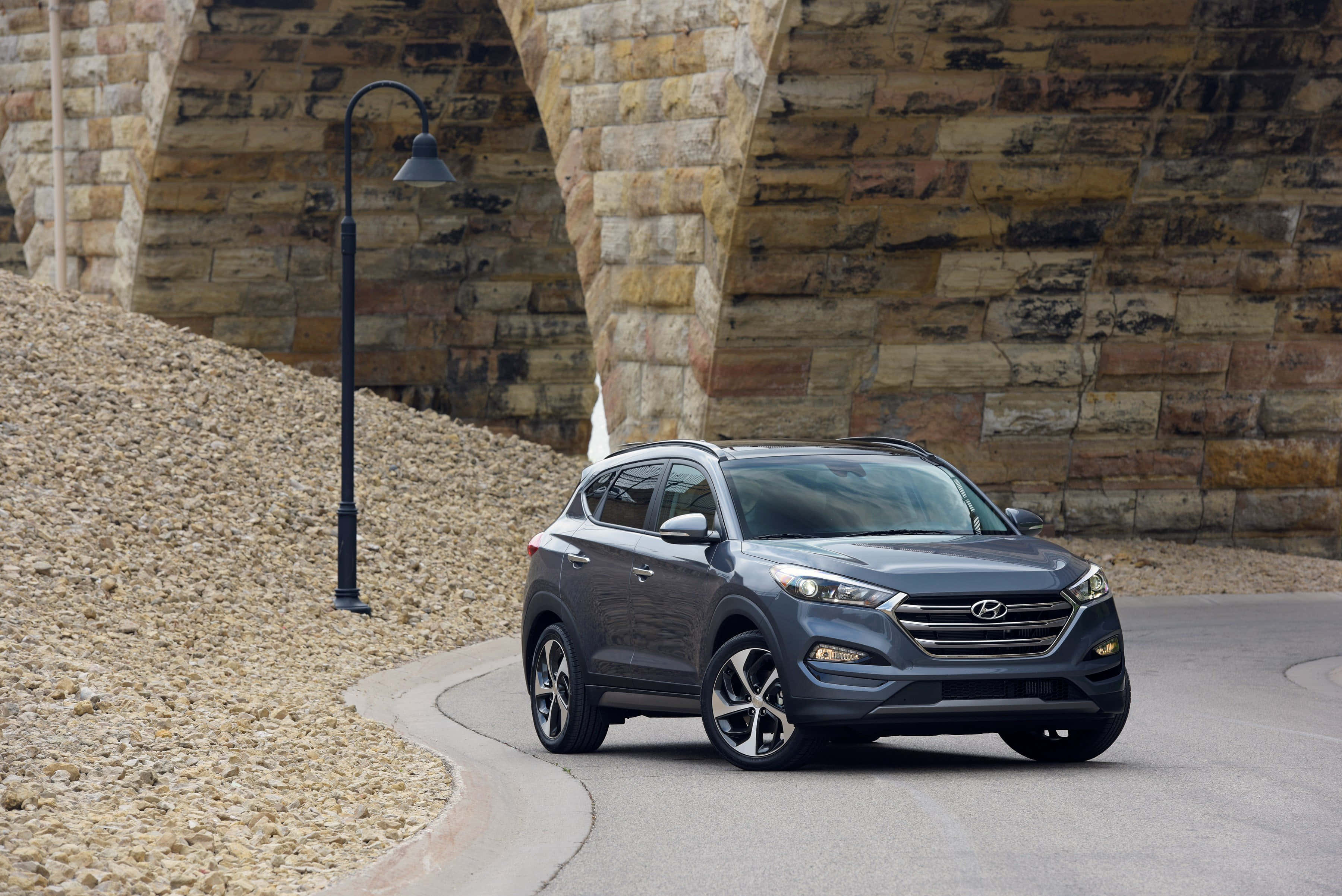 Hyundai Tucson 桌布 壁紙 — 一輛時尚的灰色Hyundai Tucson SUV在碎石河岸的質樸石橋下行駛於彎曲的道路上。