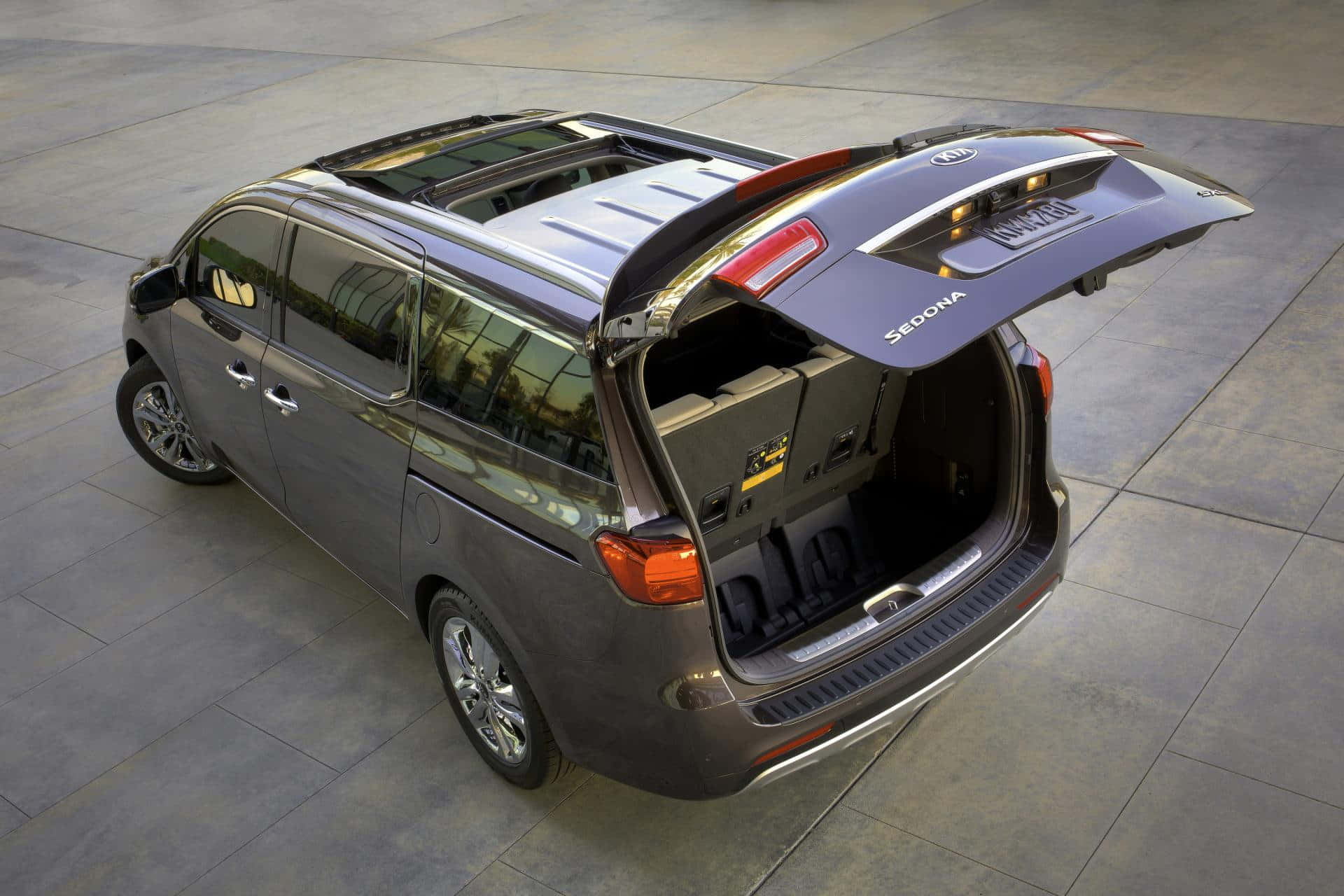 Kia Sedona 桌布 壁紙 — 一架時尚的灰色Kia Sedona小型貨車，尾門打開，露出舖磚路面上的寬敞貨物空間。