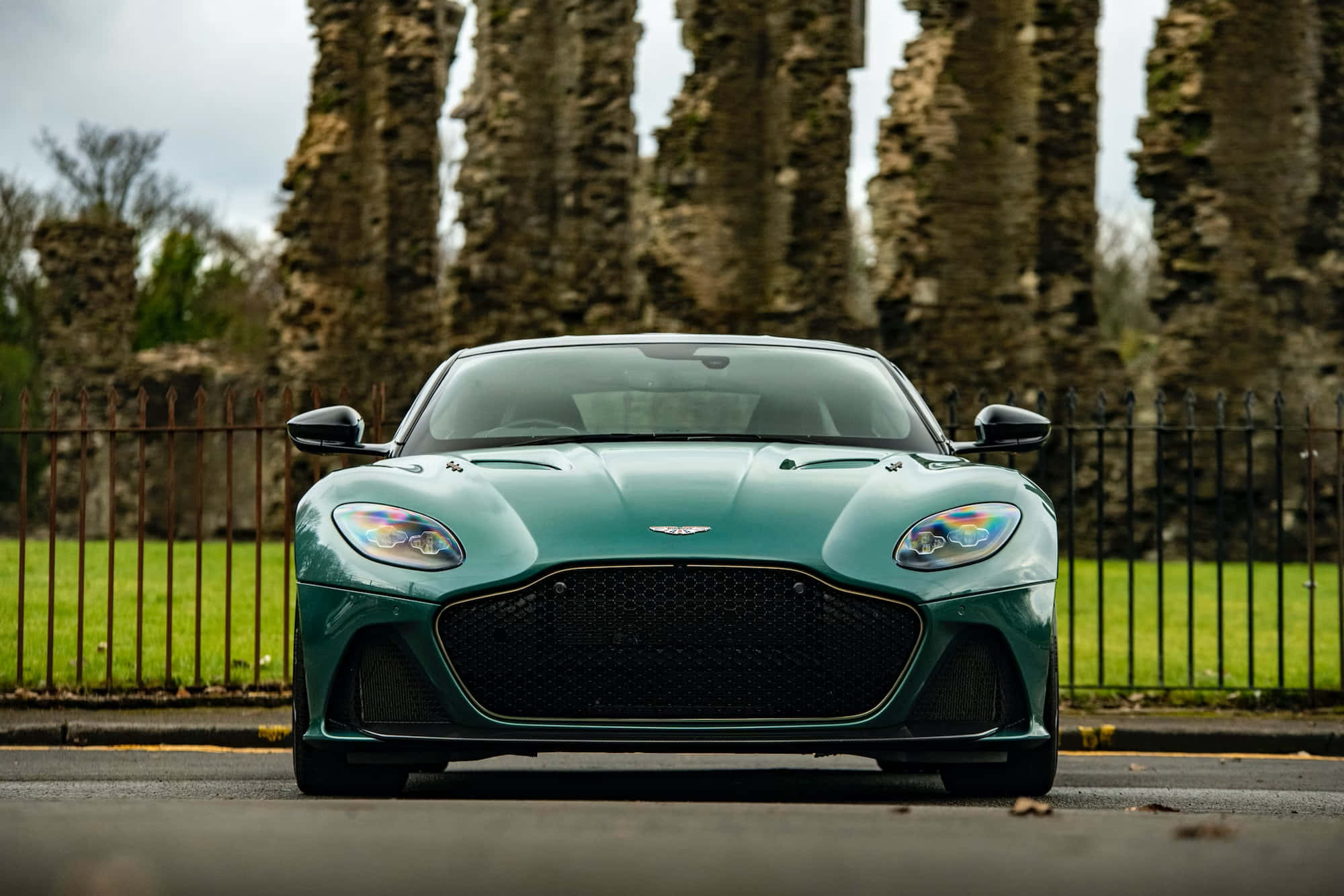 Aston Martin Dbs Superleggera 桌布 壁紙 — 一輛時尚的綠色 Aston Martin 跑車面向前方，背後有古老的石頭遺跡高聳入雲。