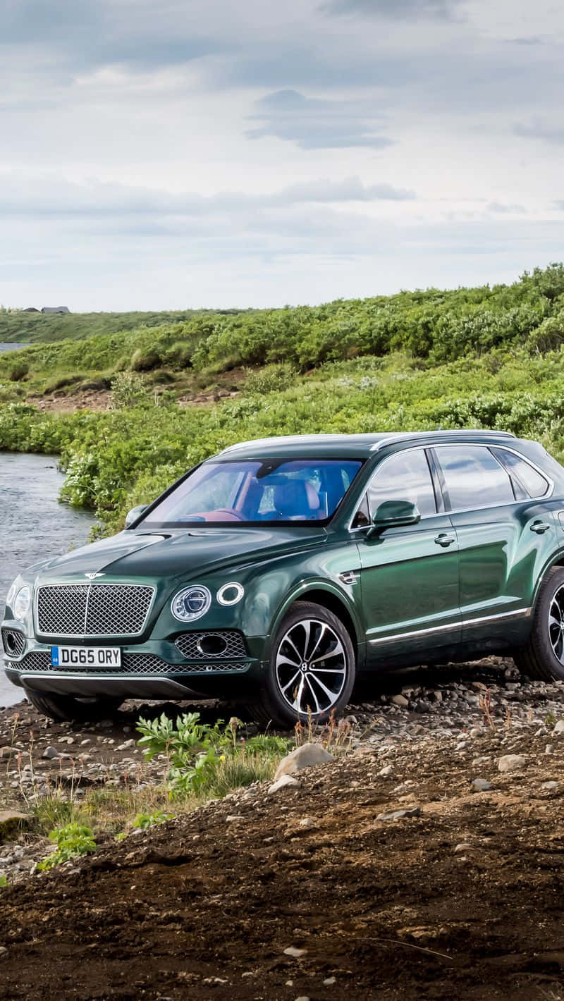Bentley Bentayga 桌布 壁紙 — 一架時尚的綠色賓利Bentayga SUV停泊在岩石河邊，背景是茂密的綠色山丘，天空多雲。