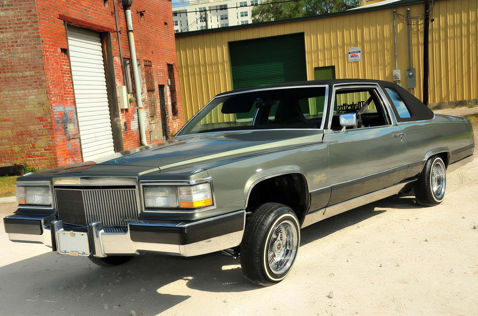 Cadillac Deville 桌布 壁紙 — 一輛時尚的綠色經典肌肉車，配備鍍鉻輪圈，停在紅磚和黃色金屬建築前，在明亮的日光下。