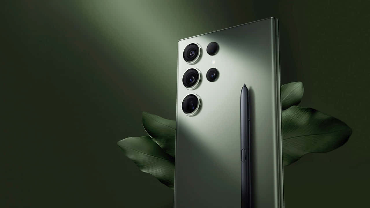 Samsung S23 Ultra 桌布 壁紙 — 一部時尚的綠色智能手機配備觸控筆，在柔和定向燈光下直立，周圍環繞著大片深色葉子。
