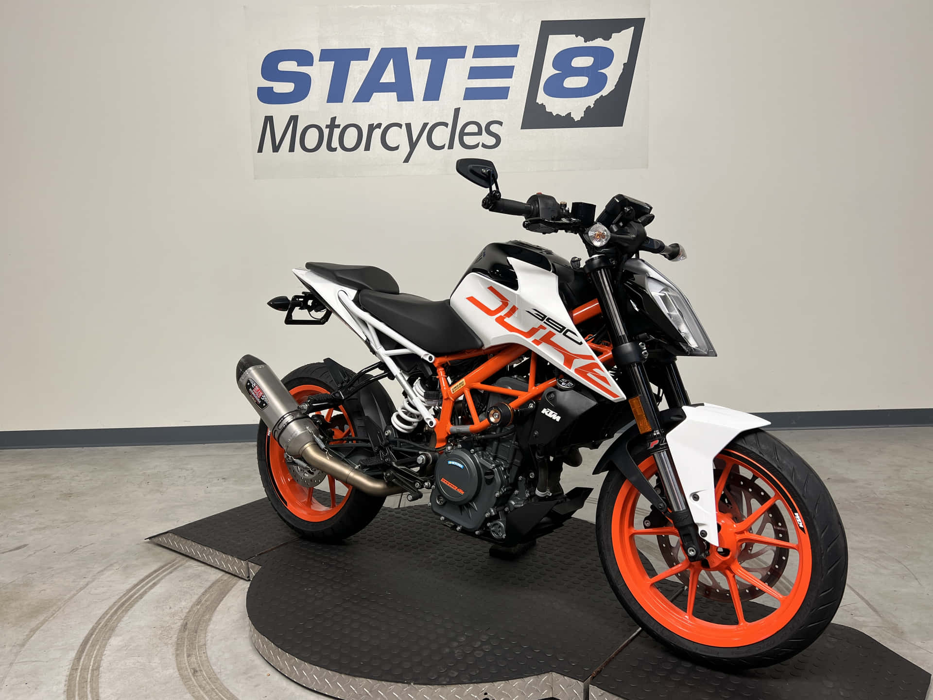 KTM Duke 390 於 State 8 Motorcycles 店前展示 壁紙 — 一輛時尚的KTM Duke電單車，帶有橙色和白色點綴，停放在展廳的黑色平台上，牆上掛有State 8 Motorcycles標誌。