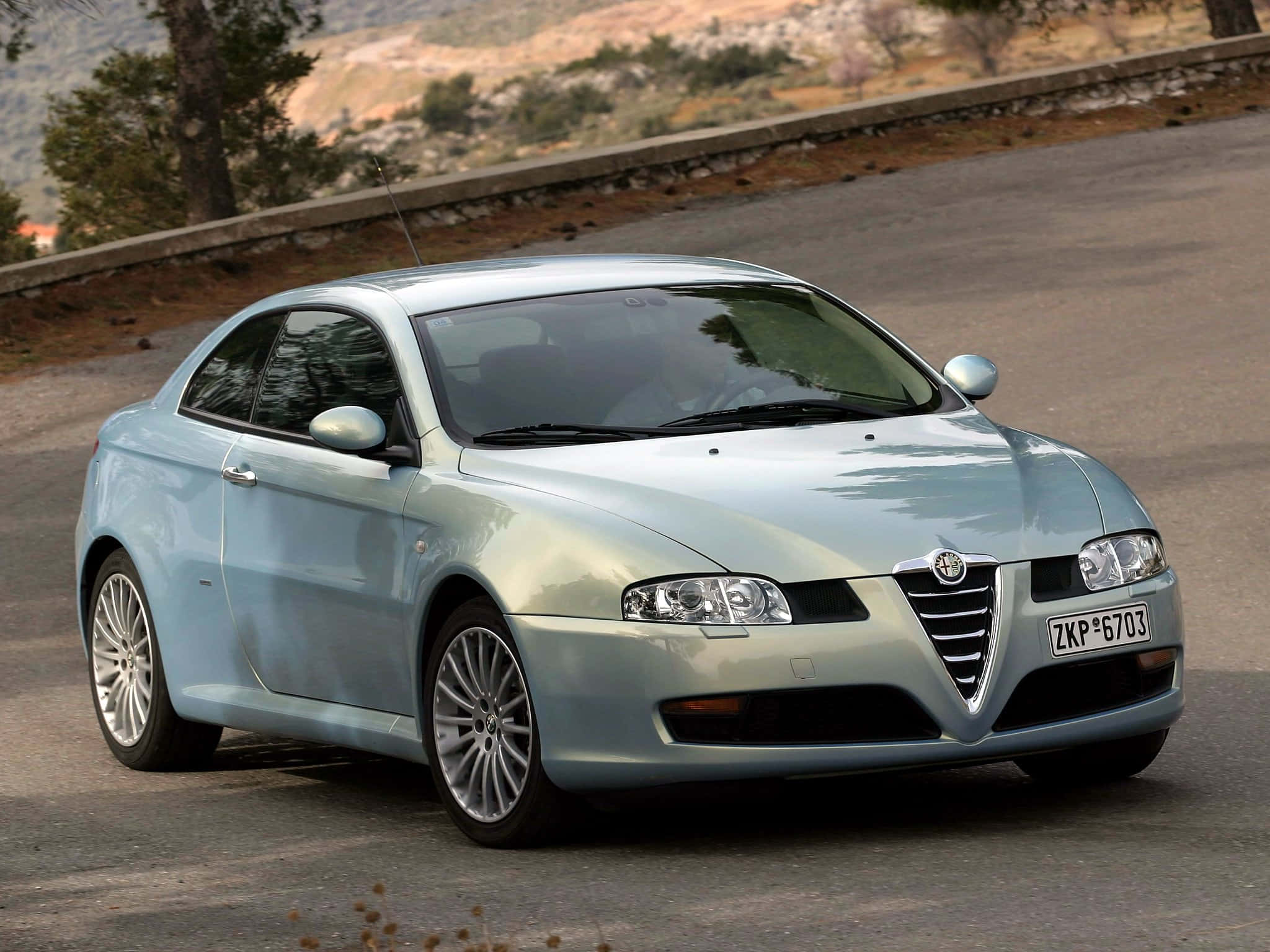 Alfa Romeo Gt 桌布 壁紙 — 一輛時尚的淺藍色愛快羅密歐轎跑車在明亮的日光下行駛在一條蜿蜒的山路上，兩旁有乾燥的植被和石頭護欄。