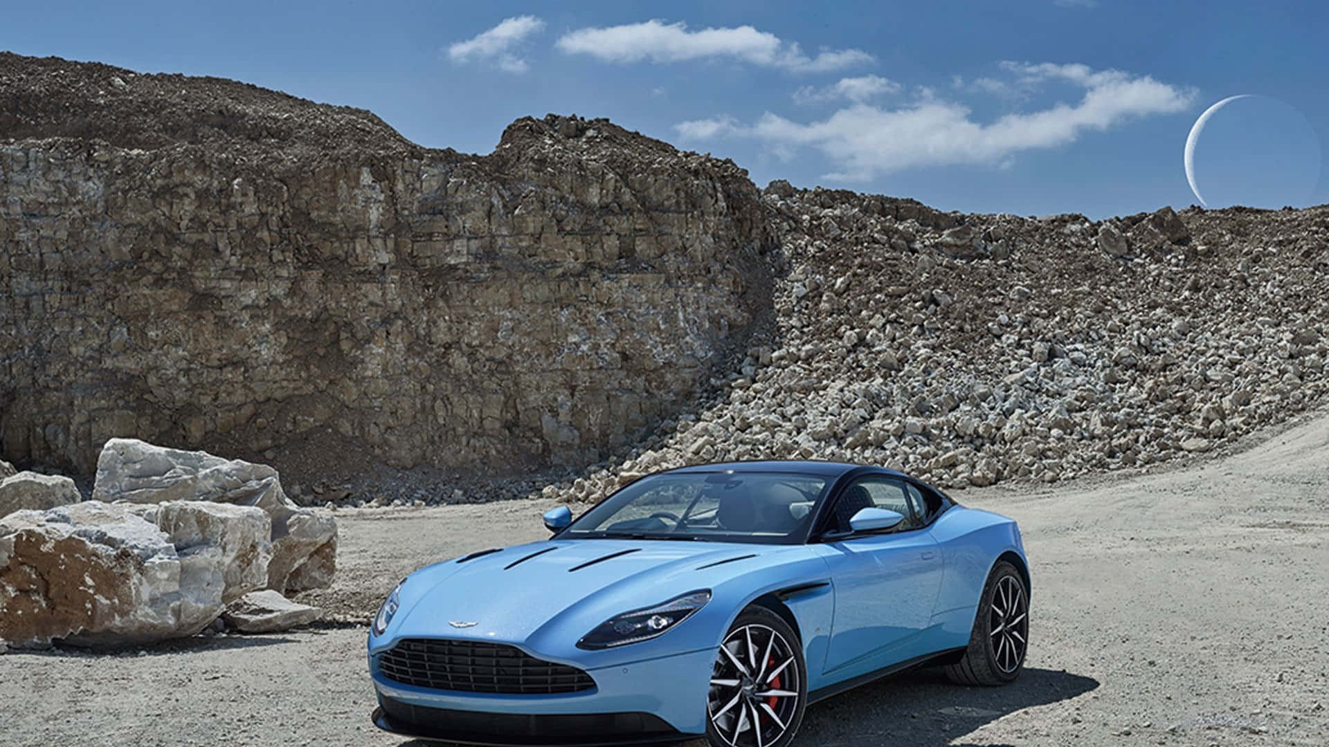 Aston Martin Db11 桌布 壁紙 — 一架時尚的淺藍色Aston Martin跑車停泊在碎石路上，旁邊是岩石懸崖，上方是明亮的藍天，掛著一彎新月。