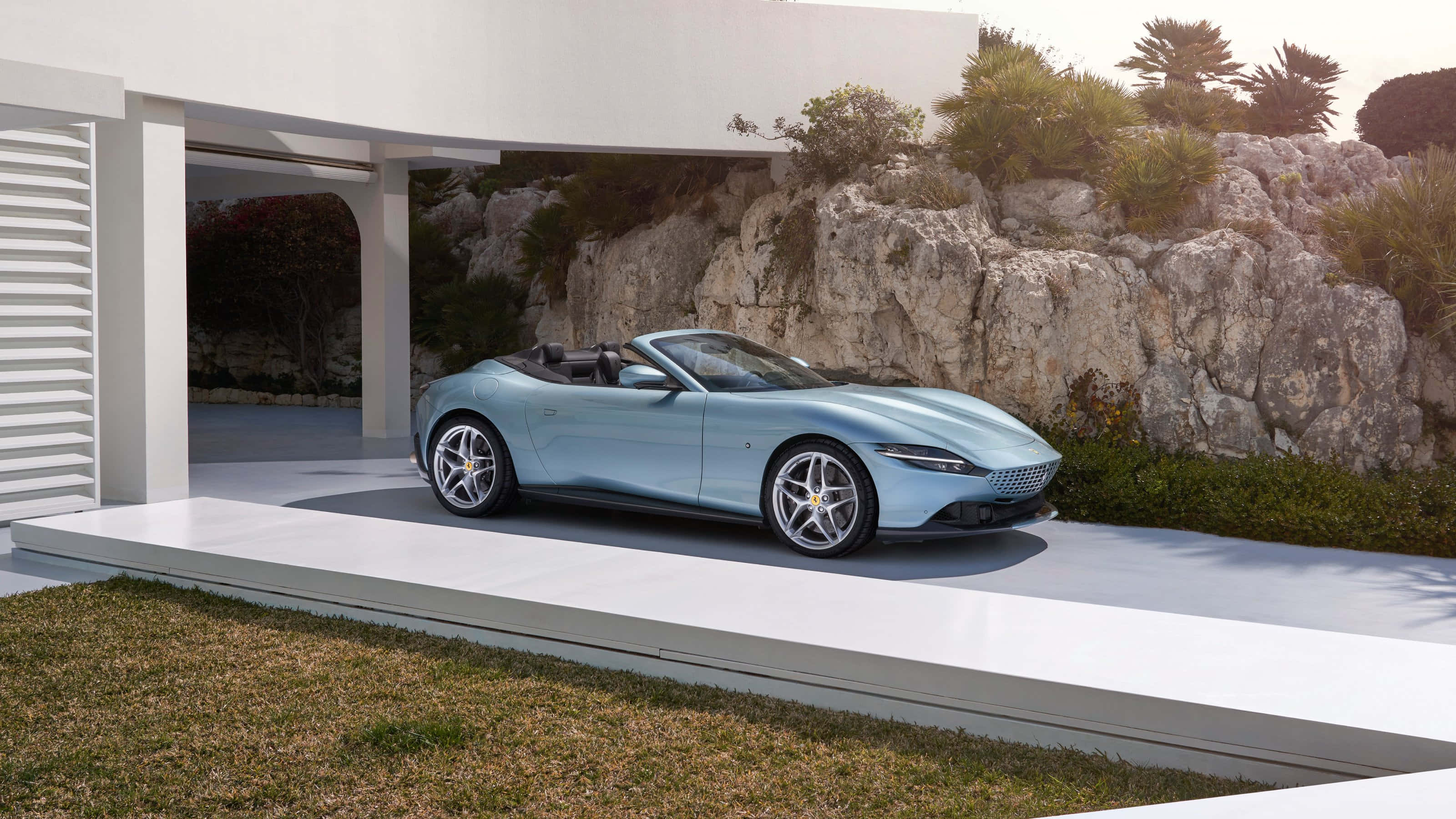 Ferrari Roma 桌布 壁紙 — 一架時尚淺藍色開篷跑車停泊在現代白色平台上，旁邊是簡約風格的房屋，背景有岩石地形和綠植。
