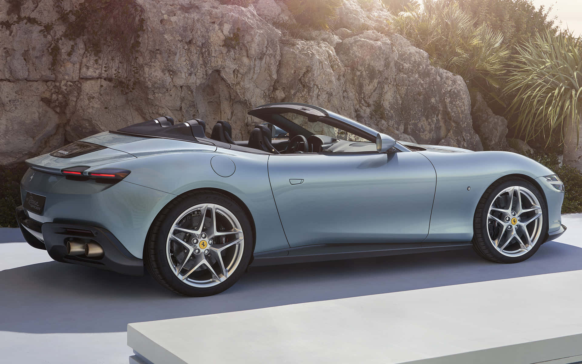 Ferrari Roma 桌布 壁紙 — 一架時尚的淺藍色法拉利開篷車停泊在現代化平台上，背景是陽光普照的岩石懸崖和棕櫚樹。