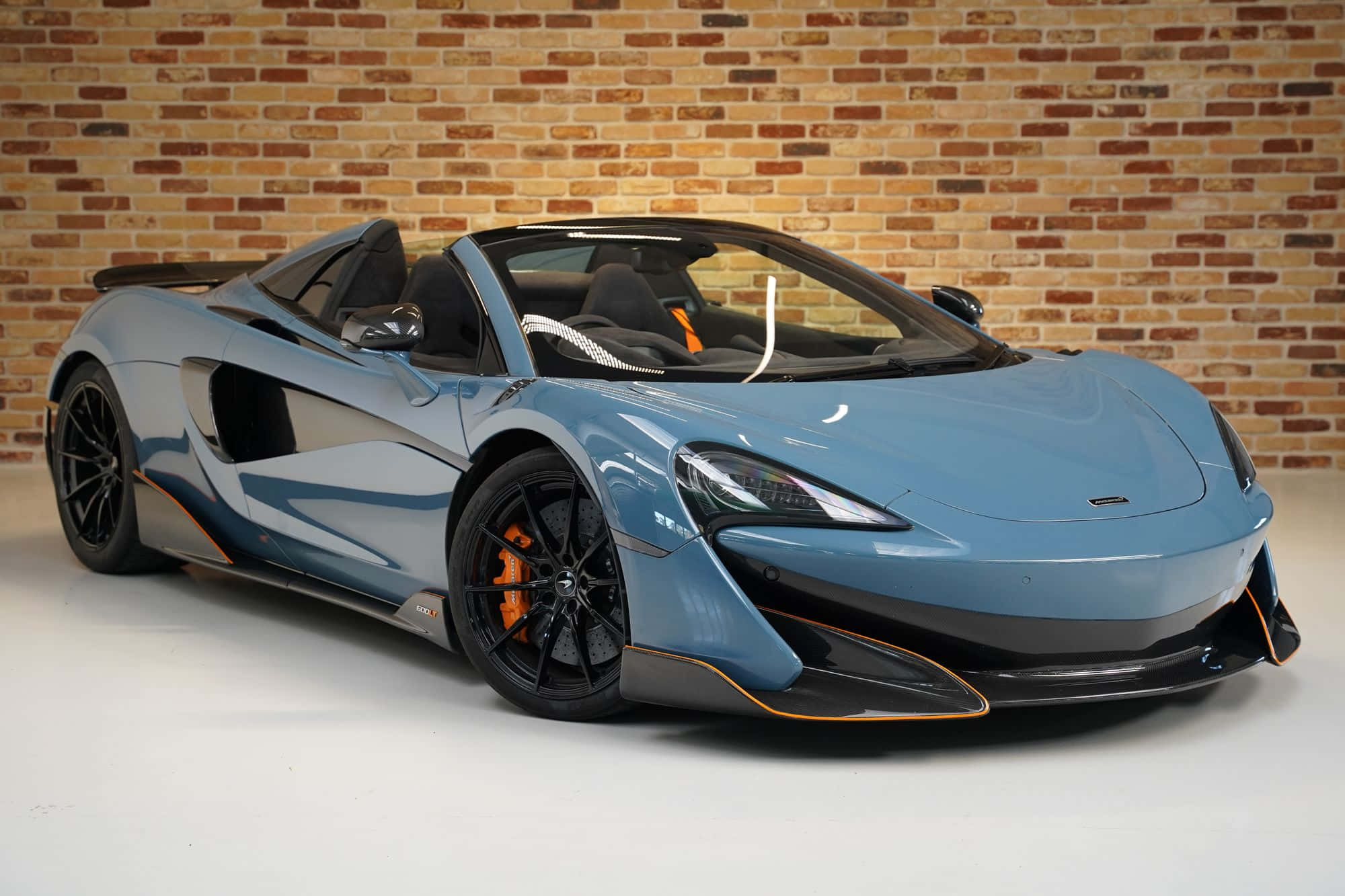 Mclaren 600lt Spider 桌布 壁紙 — 一架時尚的淺藍色McLaren跑車帶有橙色點綴，停泊在白色地板上的質樸磚牆旁。