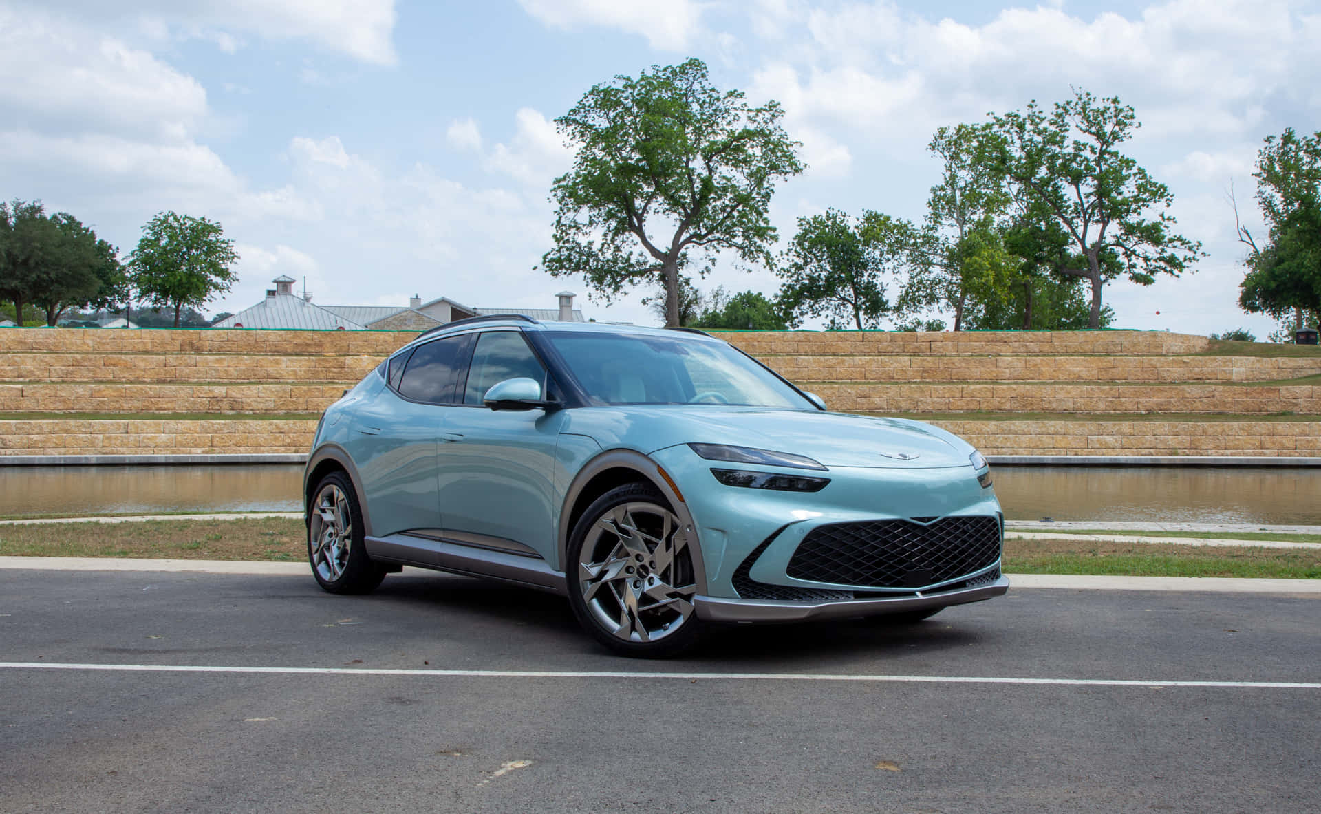 Genesis Gv60 桌布 壁紙 — 一部時尚的淺藍色現代SUV停泊在瀝青路上，旁邊有石牆和水景，天空部分多雲。