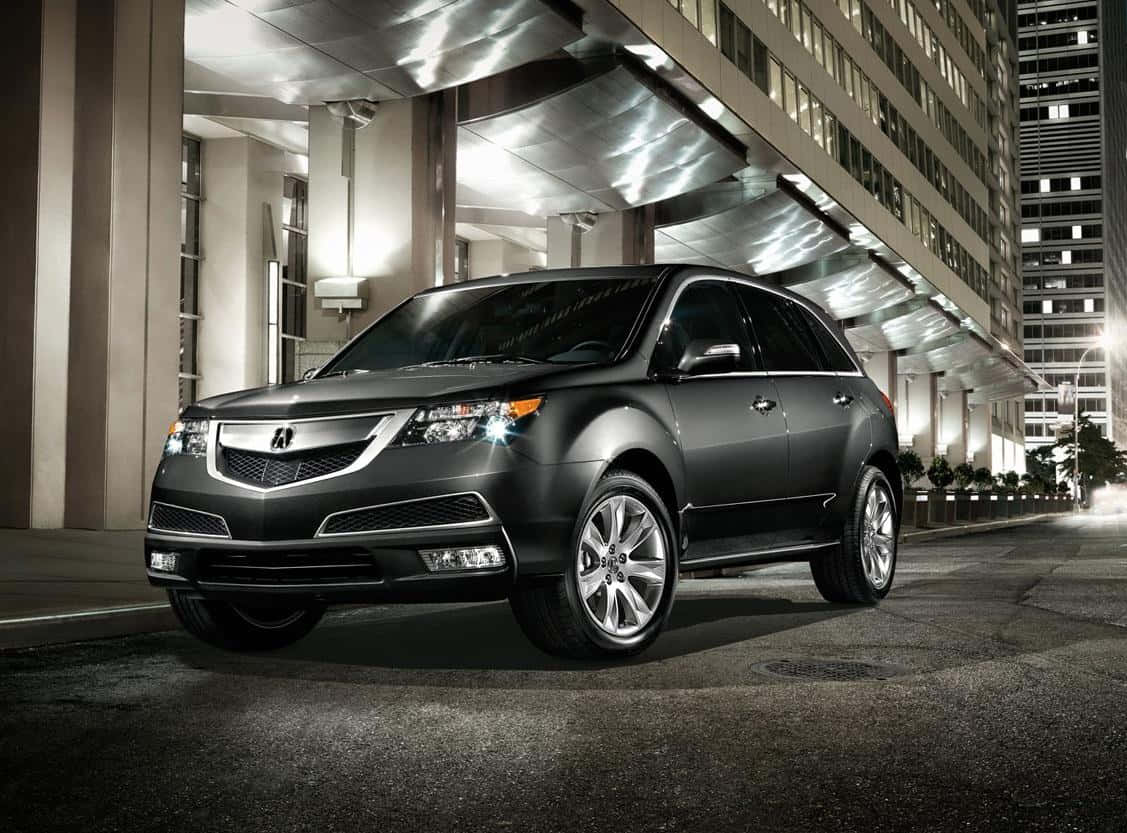 Acura Mdx 桌布 壁紙 — 一輛時尚的啞光黑色SUV停放在現代玻璃天篷下，背景是夜晚城市中燈火通明的建築物。