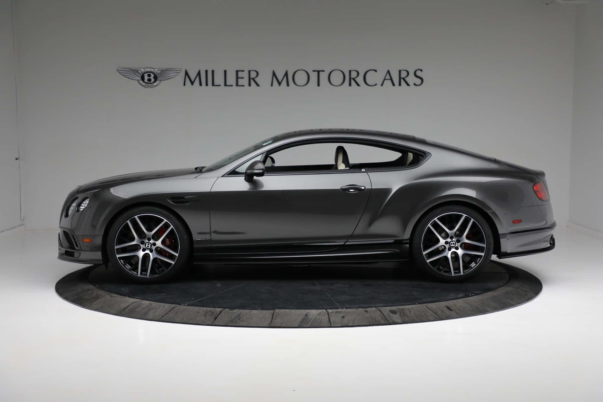 Bentley Continental Supersports 桌布 壁紙 — 一輛時尚的啞光灰色賓利 Continental GT 雙門跑車，展示在一個帶有 Miller Motorcars 品牌的簡約白色工作室的圓形平台上。