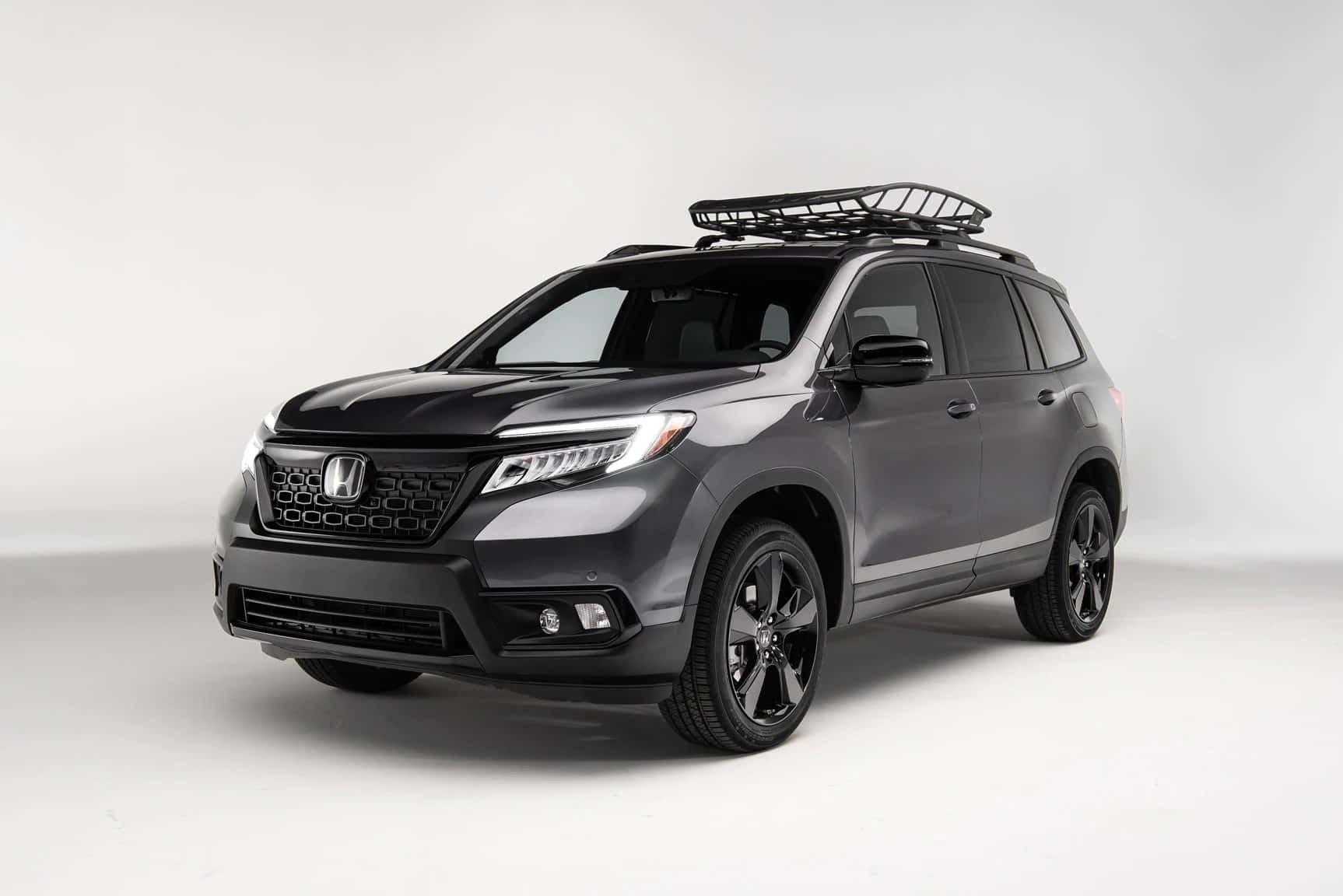 Honda Passport 桌布 壁紙 — 一部時尚的啞光灰色本田 Pilot SUV，配備黑色車頂架和車輪，擺放在乾淨的白色工作室背景前。