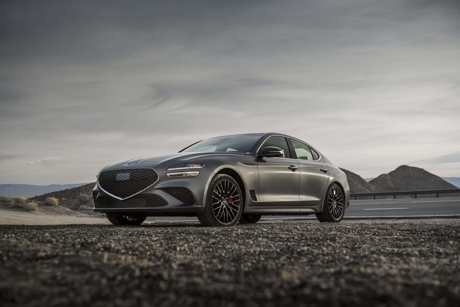Genesis G70 桌布 壁紙 — 一輛時尚的啞光灰色運動轎車配黑色輪圈，停在碎石路邊，背景是廣闊的沙漠，天空多雲。