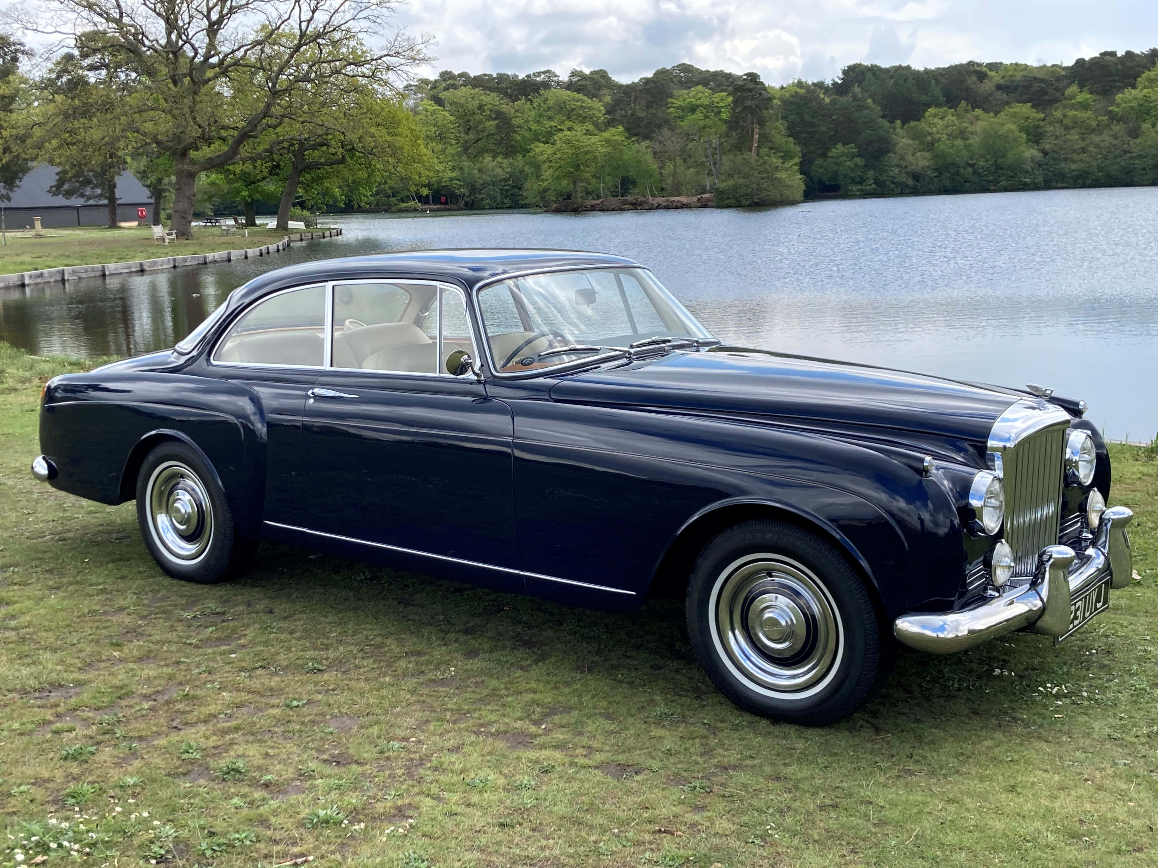 Bentley S2 桌布 壁紙 — 一架時尚的深藍色經典汽車在草地上閃閃發光，旁邊是一個寧靜的湖泊，周圍環繞著茂密的綠樹，天空部分多雲。