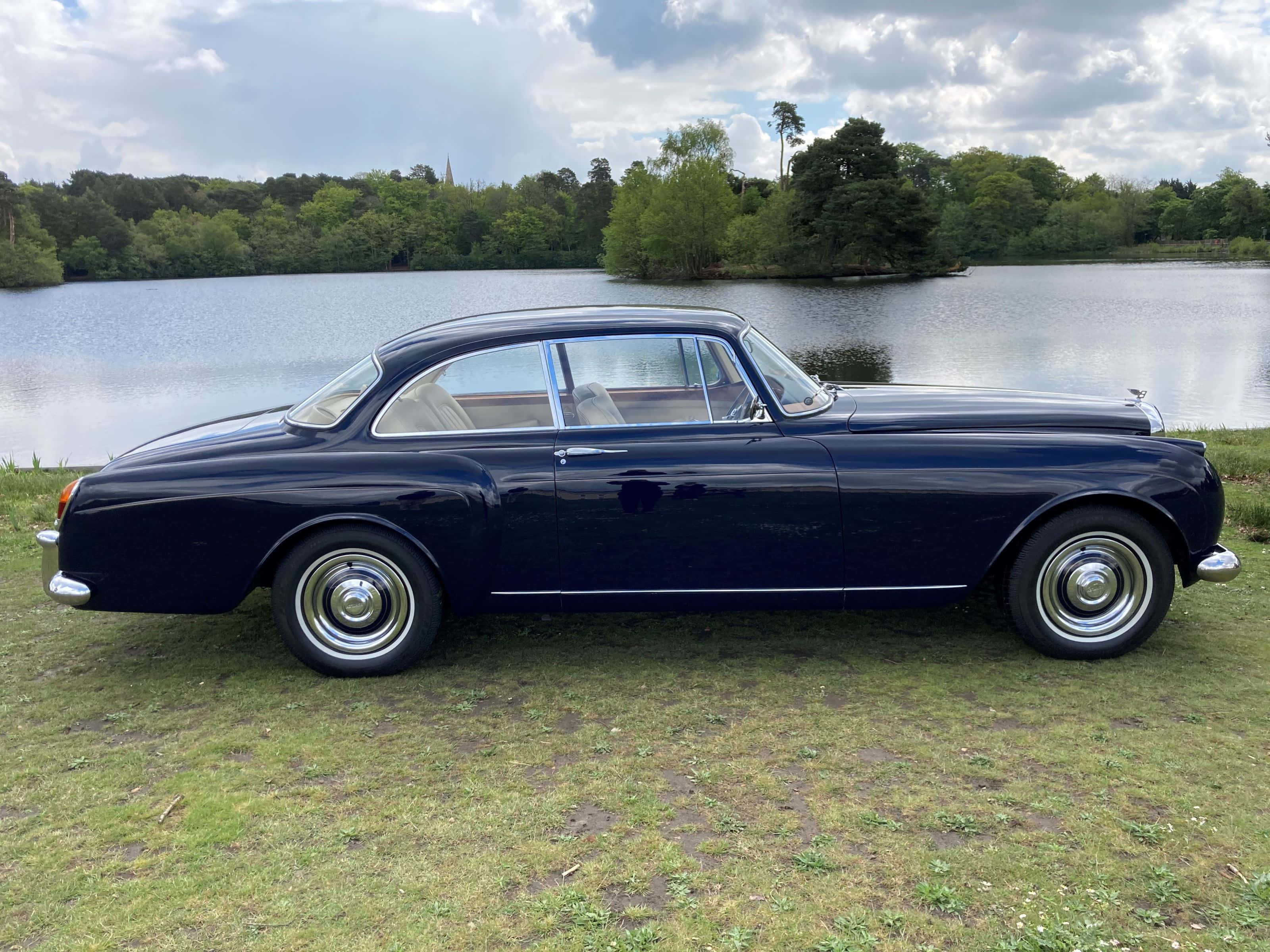 Bentley S2 桌布 壁紙 — 一部時尚的深藍色經典汽車停在草地上，旁邊是一個平靜的湖泊，周圍環繞著茂密的綠樹，天空部分多雲。